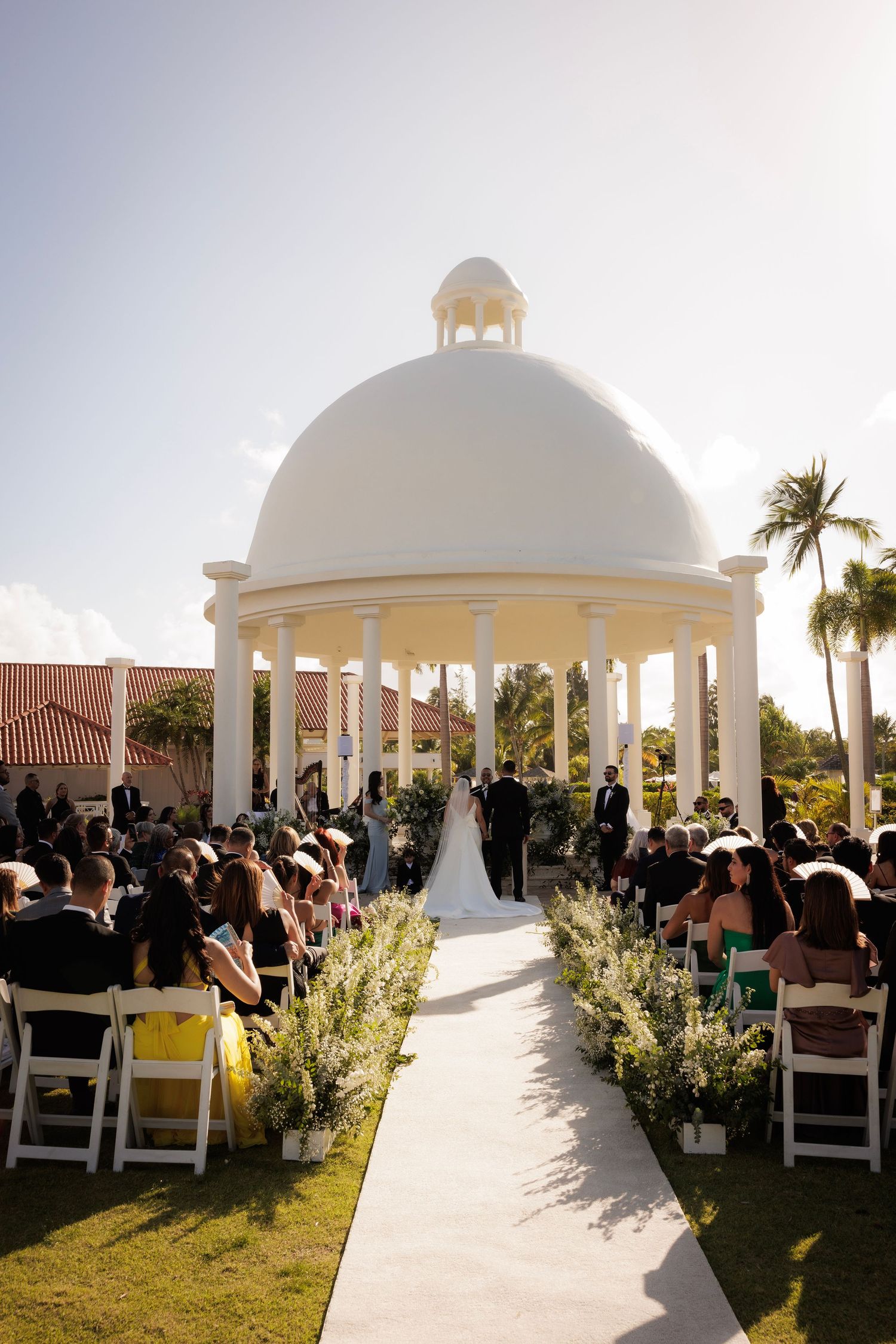 Elegant outdoor wedding ceremony under white domed gazebos with guests seated on white chairs against a sunset backdrop.