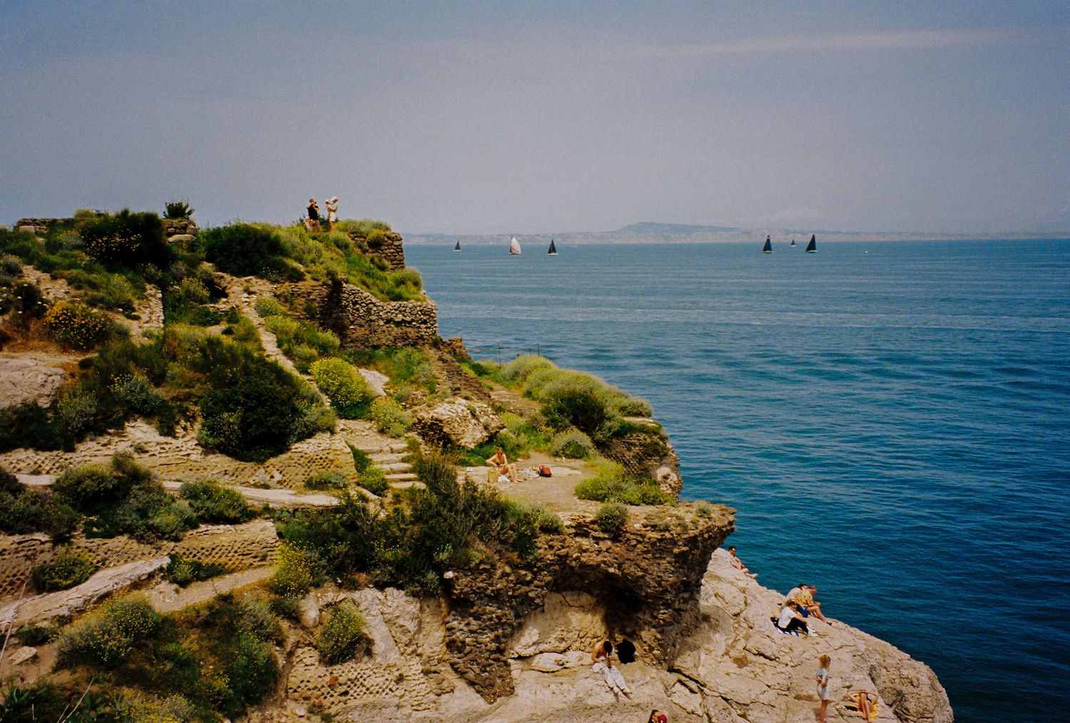 Rocky coastal cliffs overlooking calm blue Mediterranean sea with sailboats and lush green vegetation under hazy sky.