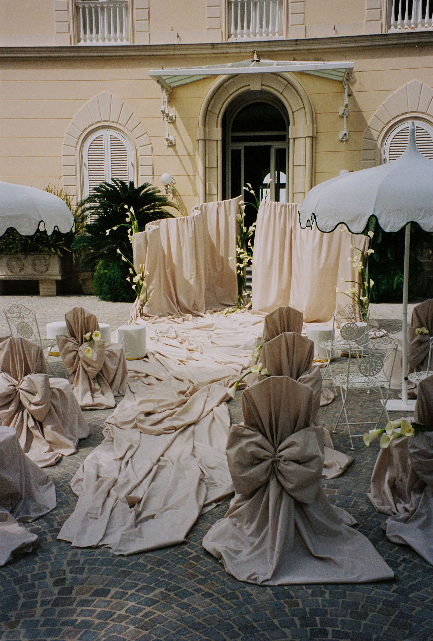 Elegant outdoor wedding ceremony setup with draped fabric chairs, floral accents, and white umbrellas in a courtyard.