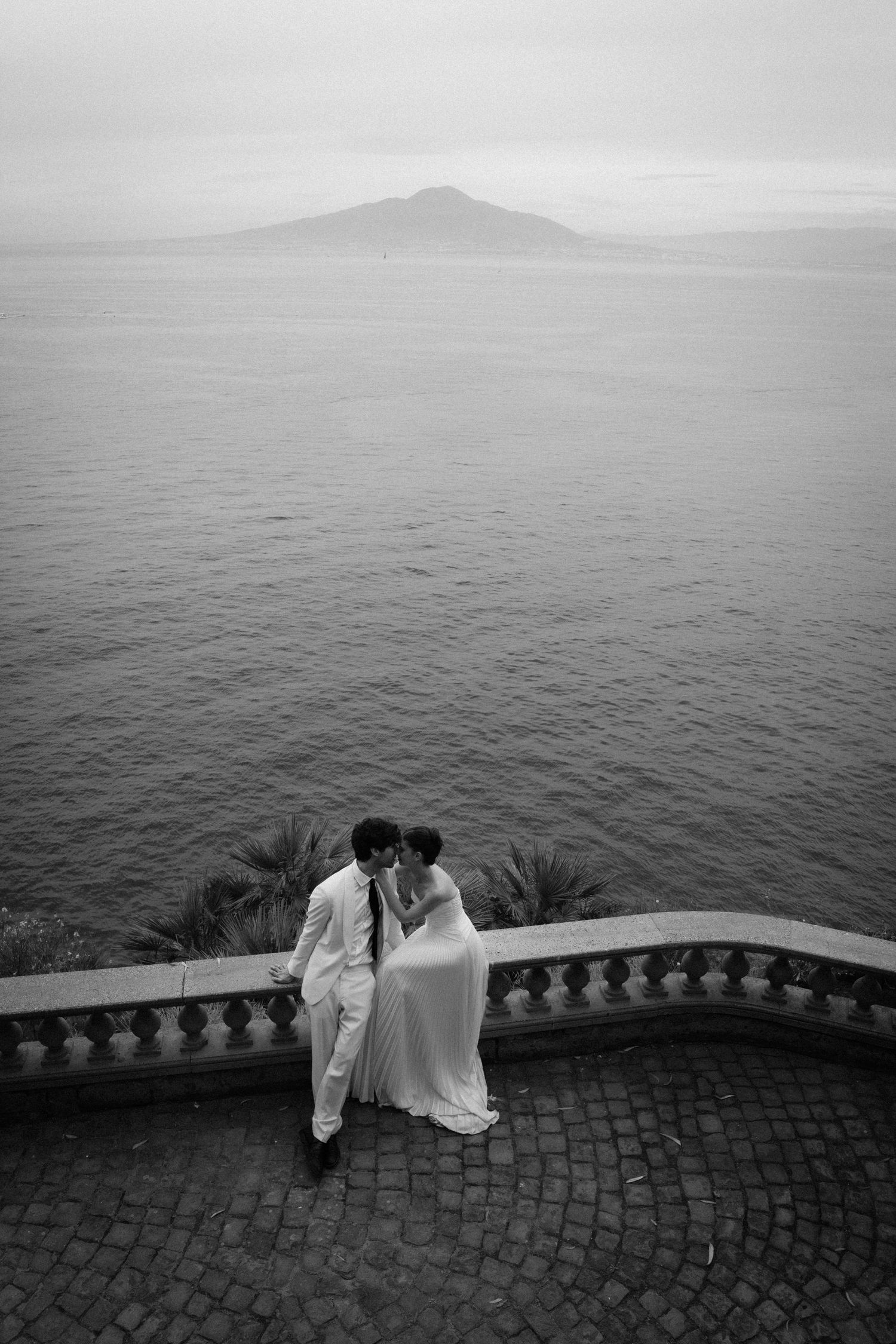 Black and white photo of a couple in wedding attire sharing a romantic moment on a terrace overlooking the sea.
