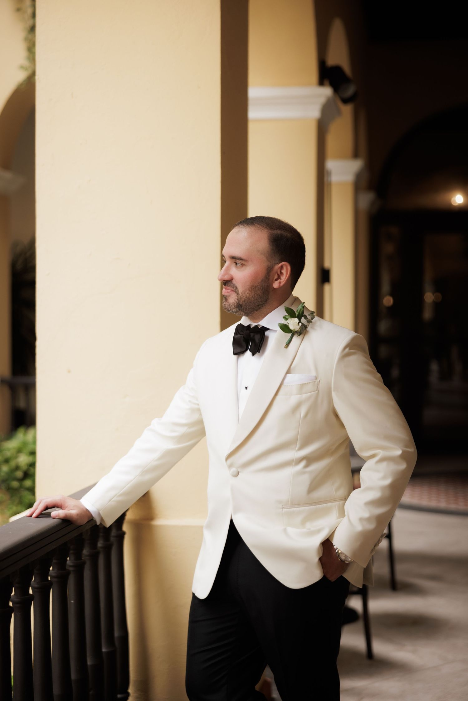 A person in a white dinner jacket poses against columns in an elegant outdoor corridor.