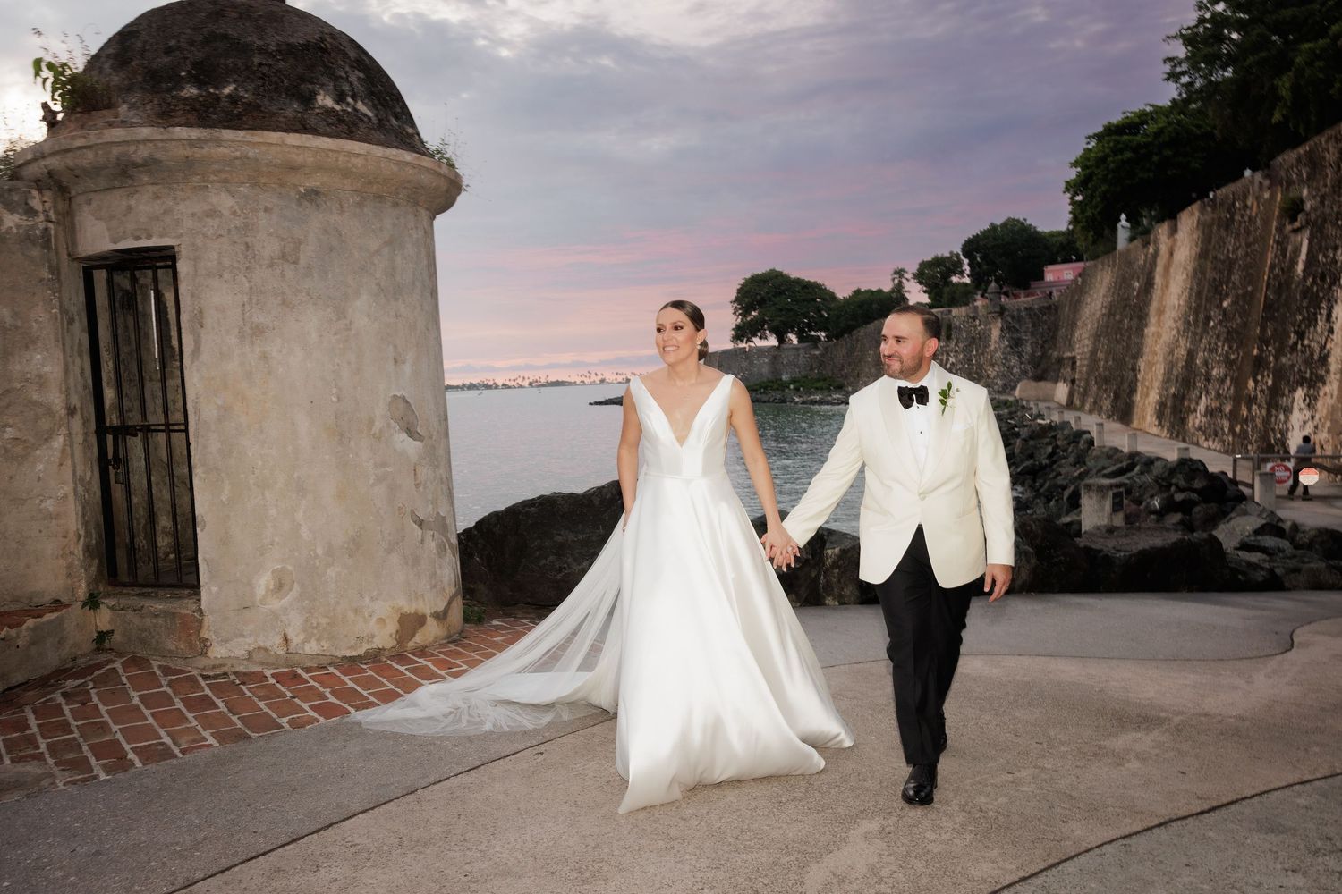 Wedding couple poses against stone walls and scenic coastal views at sunset.