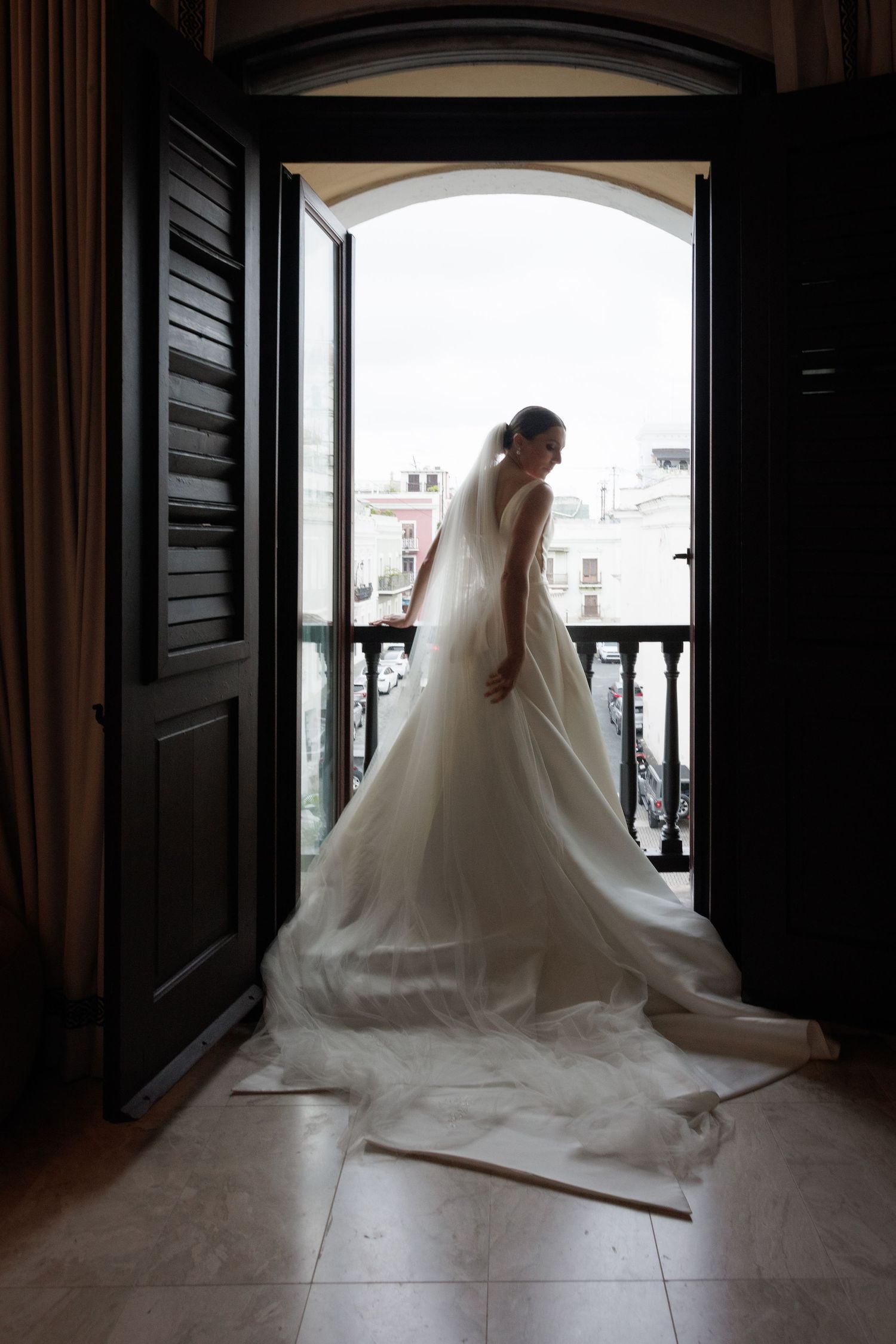 A bride in a flowing white wedding gown stands silhouetted against bright arched windows in an elegant interior space.