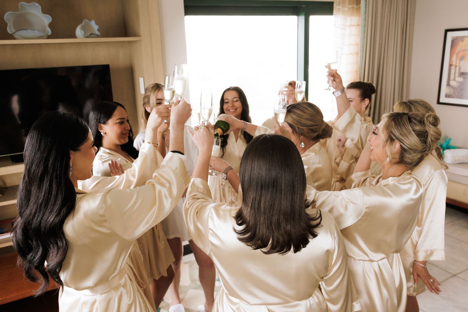 Group wearing matching cream-colored satin robes celebrating in a bright hotel room.
