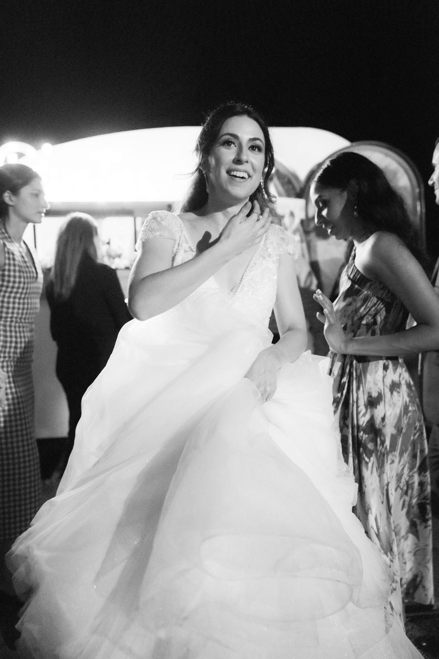 Bride in flowing white wedding gown being helped into a car with guests looking on.