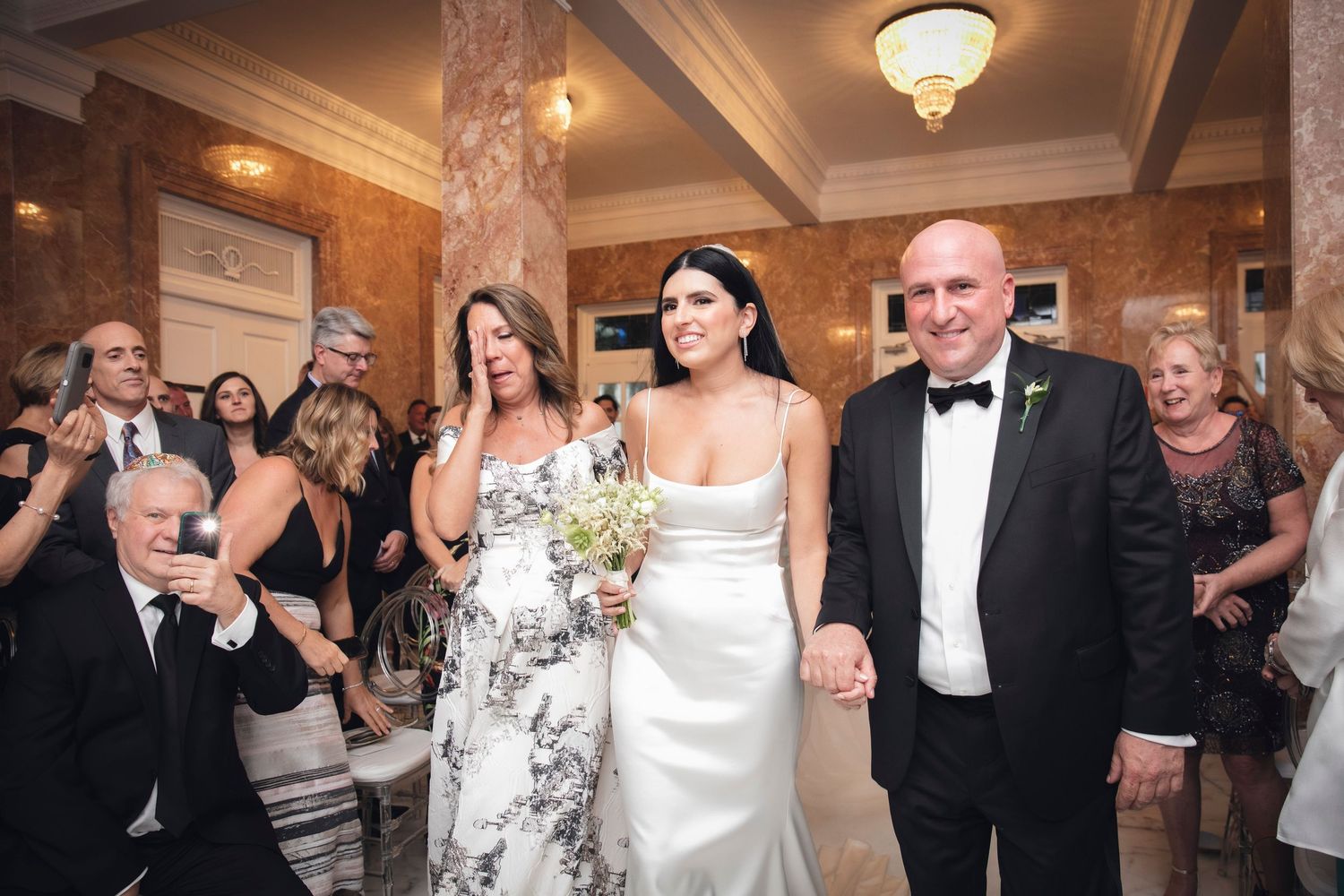 Wedding party guests dance together on the dance floor at an elegant indoor reception venue with ornate lighting.