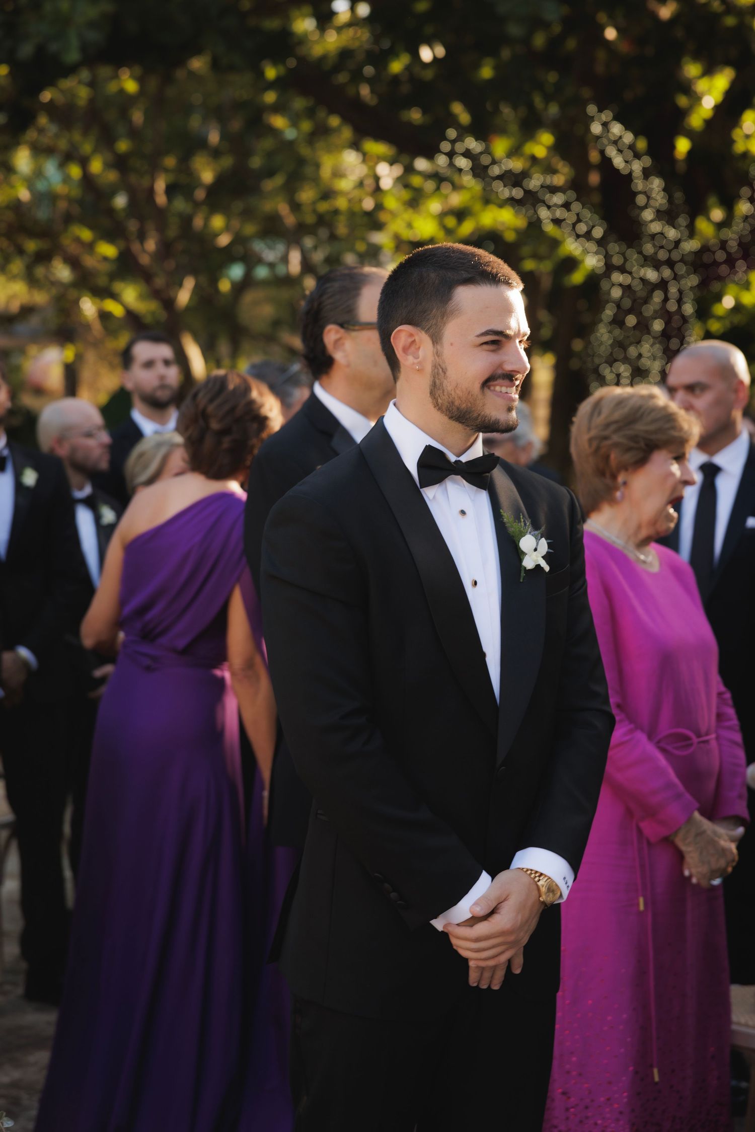 A wedding guest in a black tuxedo smiles during an outdoor ceremony with guests in purple dresses in the background.