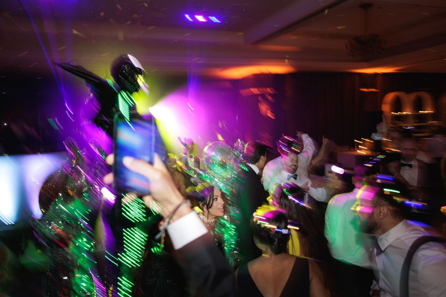 Colorful dance floor lights illuminate a crowded reception as party guests move to the music in a blur of motion.