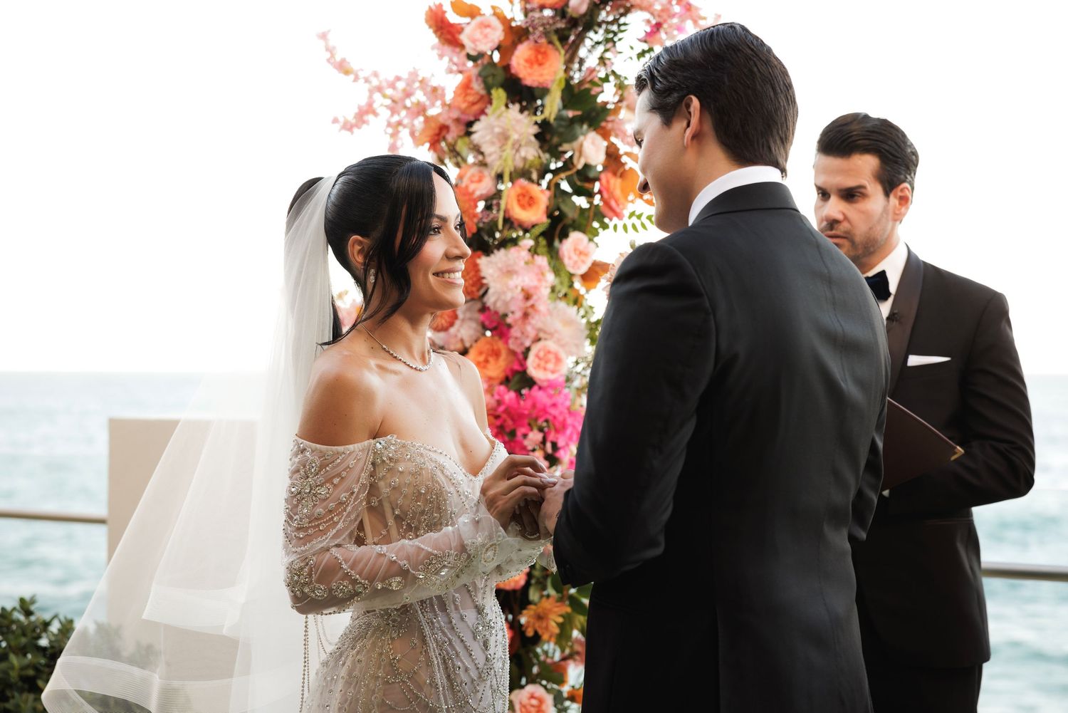 Elegant beachside wedding ceremony with floral arrangements and guests gathered under a cloudy sky.