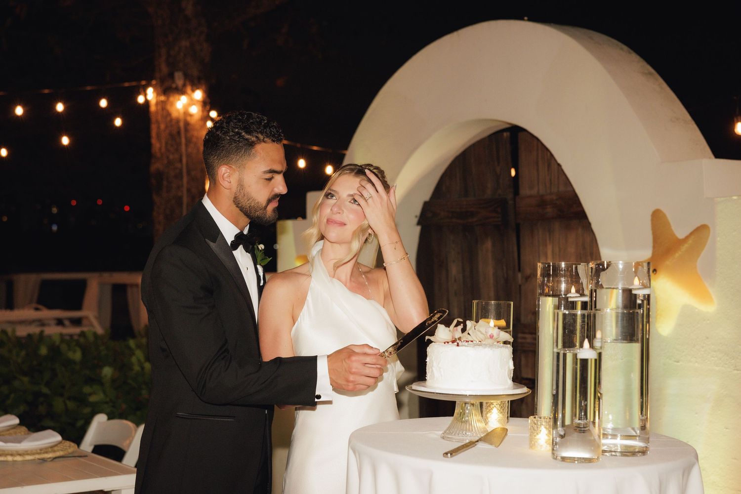 A couple shares a joyful moment while cutting their wedding cake at an outdoor reception with string lights and arches.
