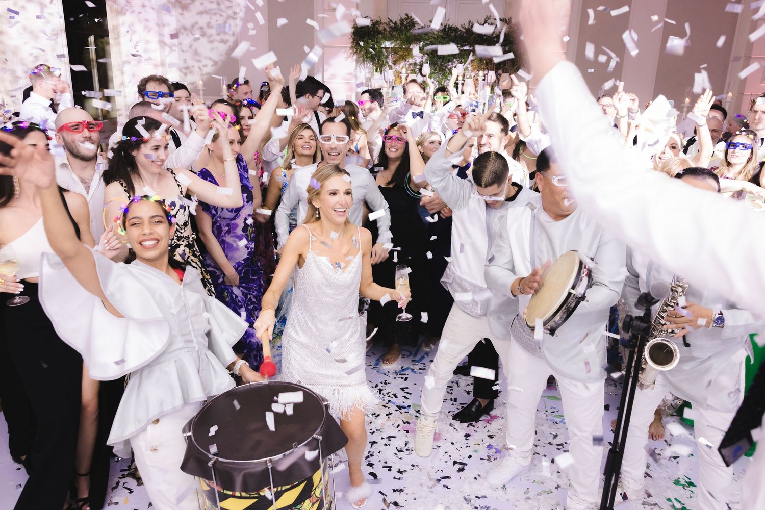 Energetic wedding reception dance party with guests celebrating under falling confetti in a white decorated venue.