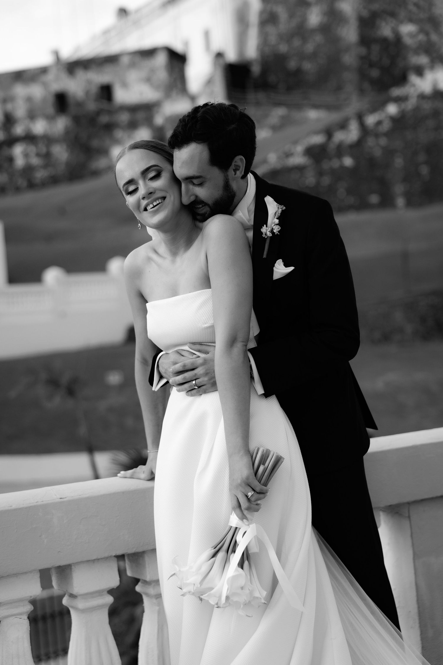 A bride and groom share an intimate black and white moment on their wedding day, leaning together against a railing.