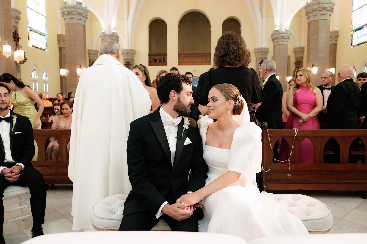 A couple smiles at each other during their wedding ceremony inside a beautiful church with ornate columns and arches.