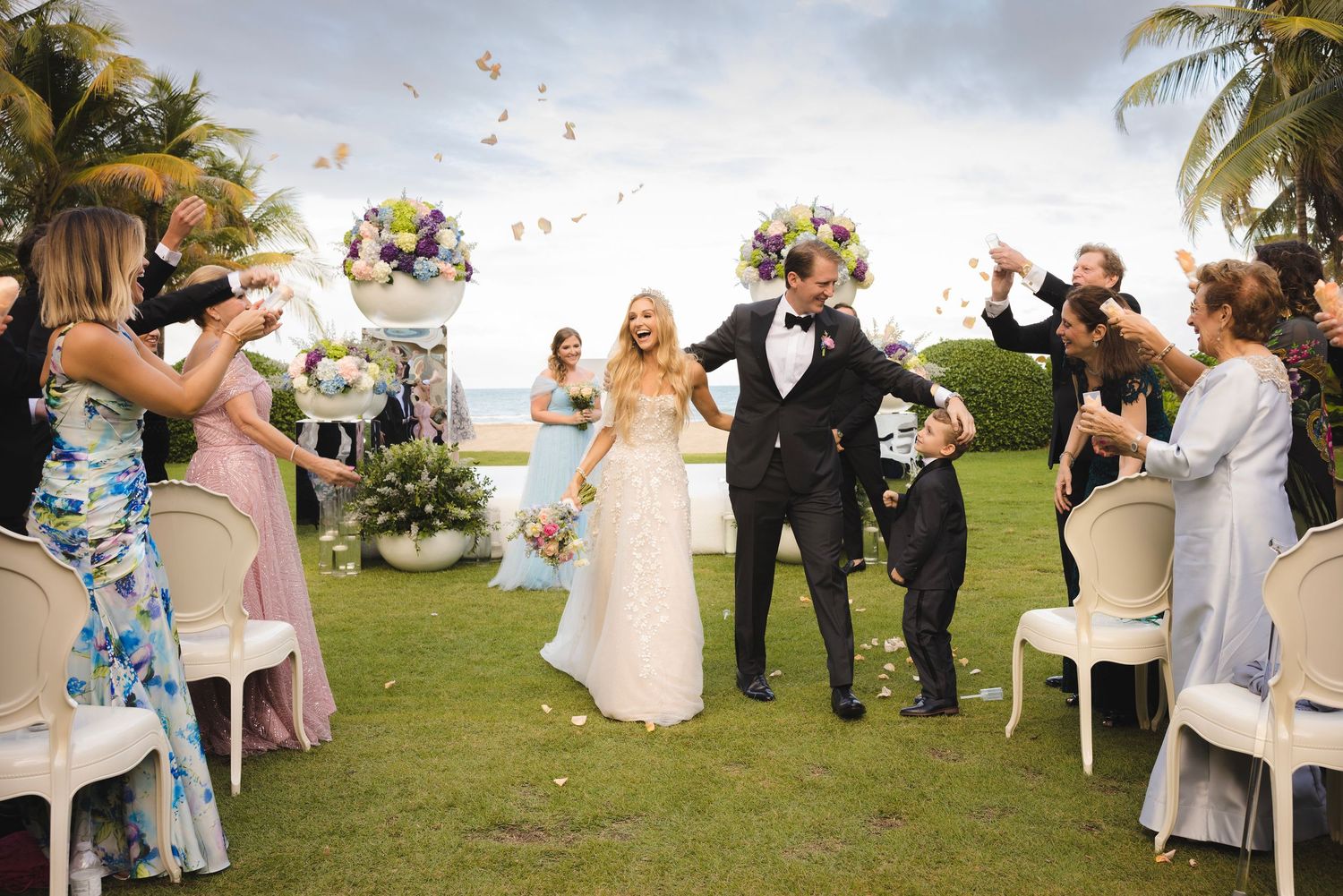 Wedding guests toss flower petals in celebration during an outdoor tropical beach ceremony.