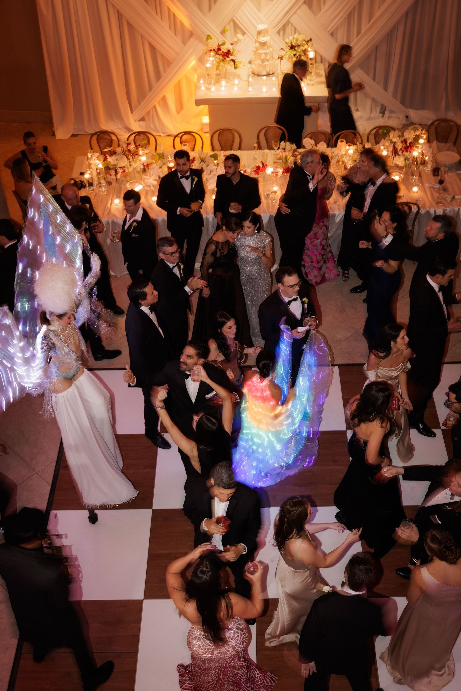 Elegant wedding reception with dancers on a black and white checkered floor illuminated by rainbow light effects.