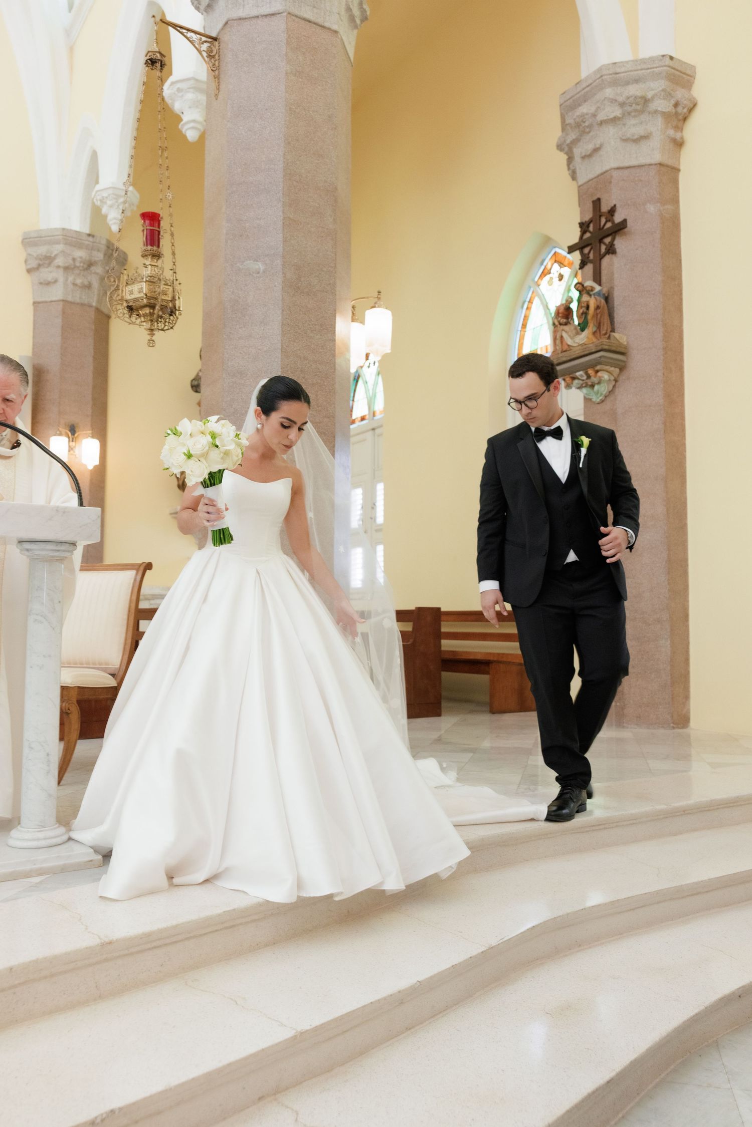 Wedding couple walks through elegant yellow-walled church corridor with arched columns.