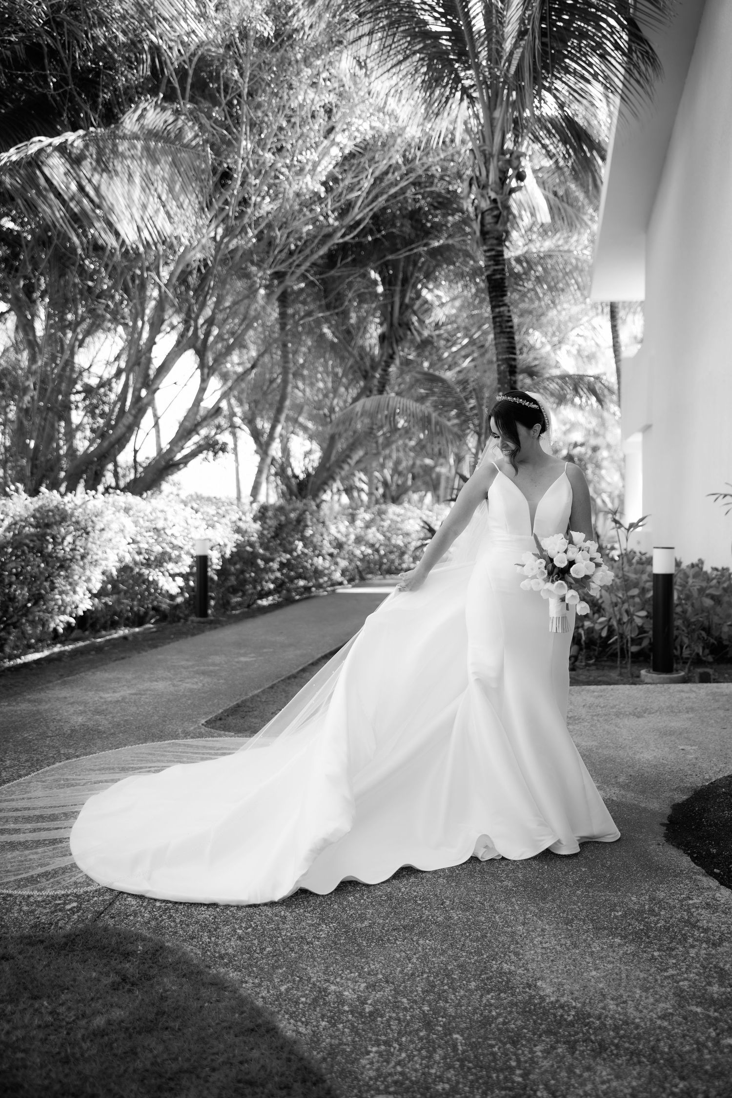 Black and white photos show a flowing white wedding dress train sweeping across a grassy path beneath tropical trees.