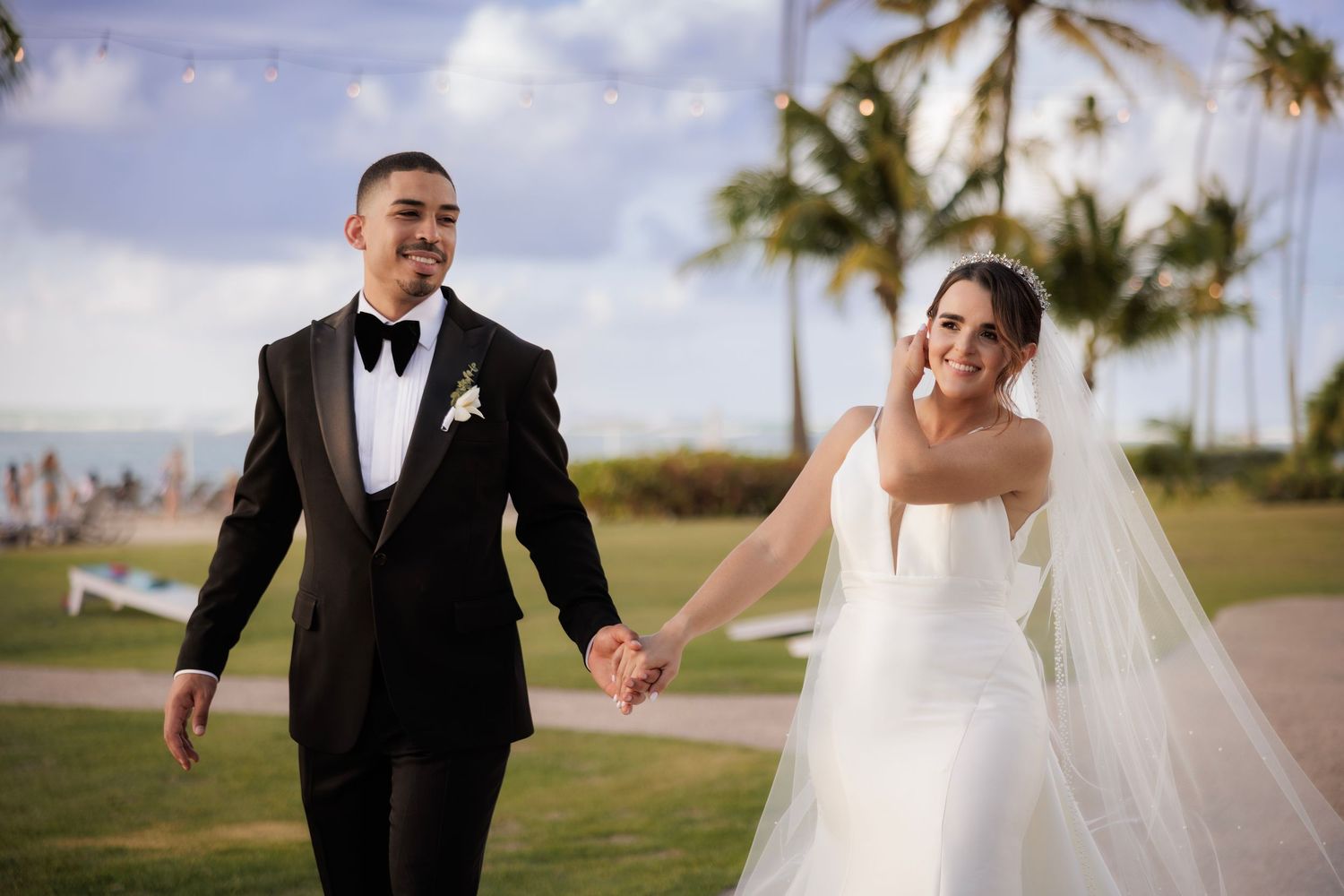 A wedding couple walks and laughs together on a palm tree-lined path by the beach during sunset.