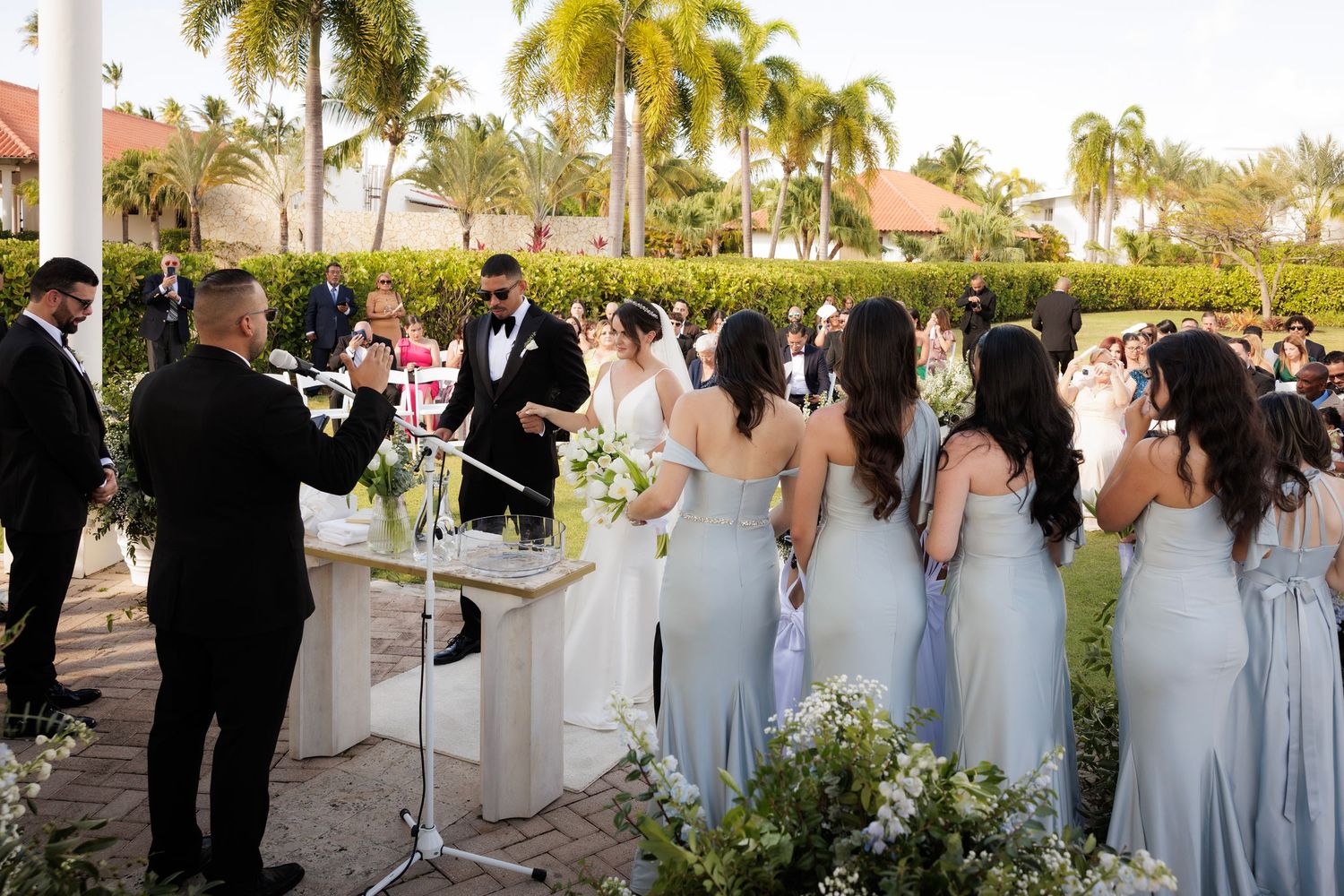 Wedding party stands in line on a palm tree-lined pathway during an elegant outdoor ceremony.