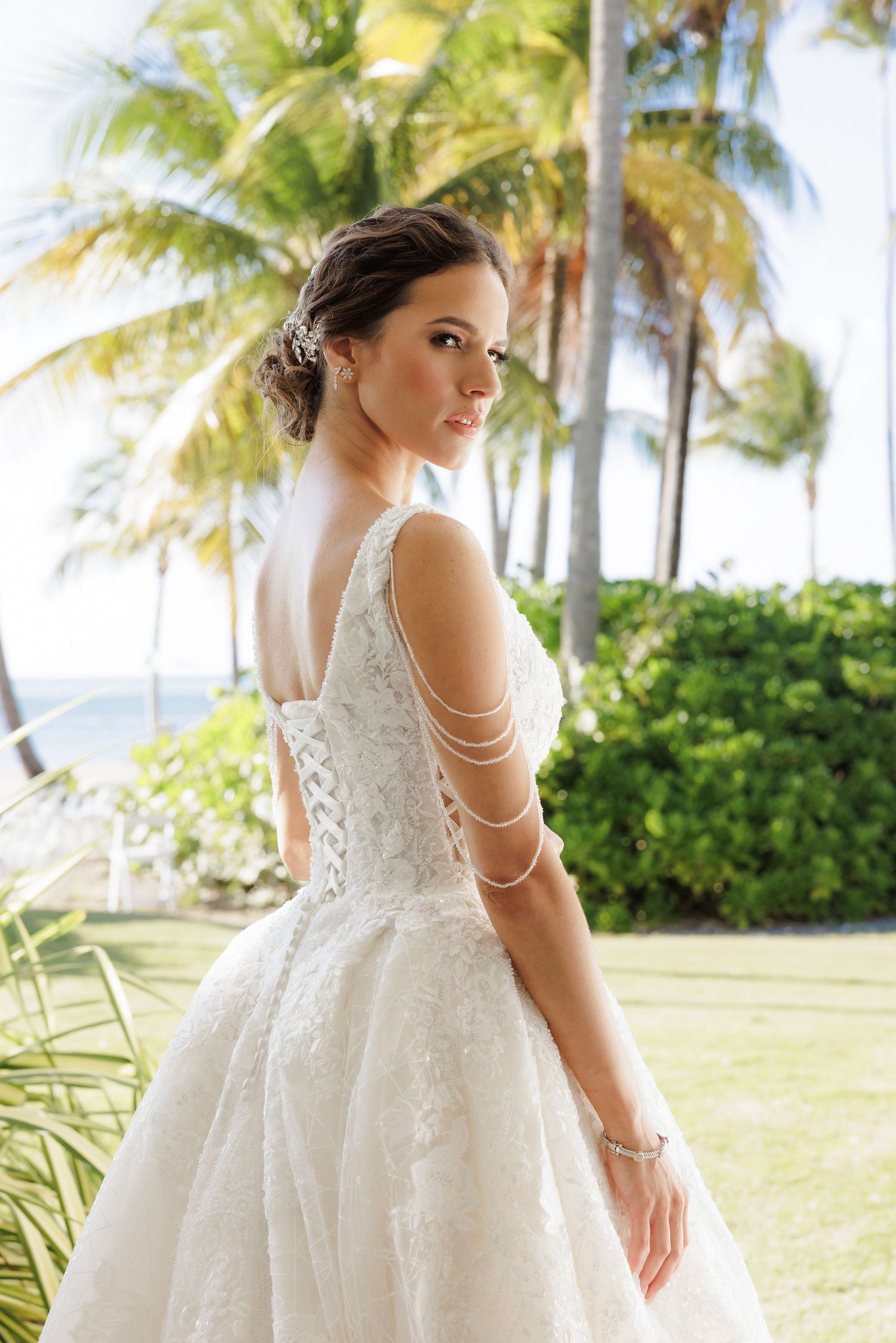 A bride in an ornate white lace gown poses elegantly on a tropical beach with palm trees and ocean in the background.