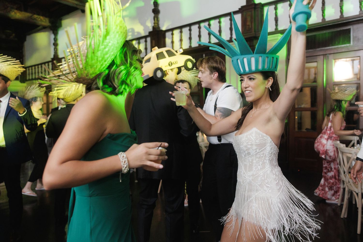 Wedding guests dance enthusiastically at a reception with someone wearing a bright green feathered headpiece and fringed dress.