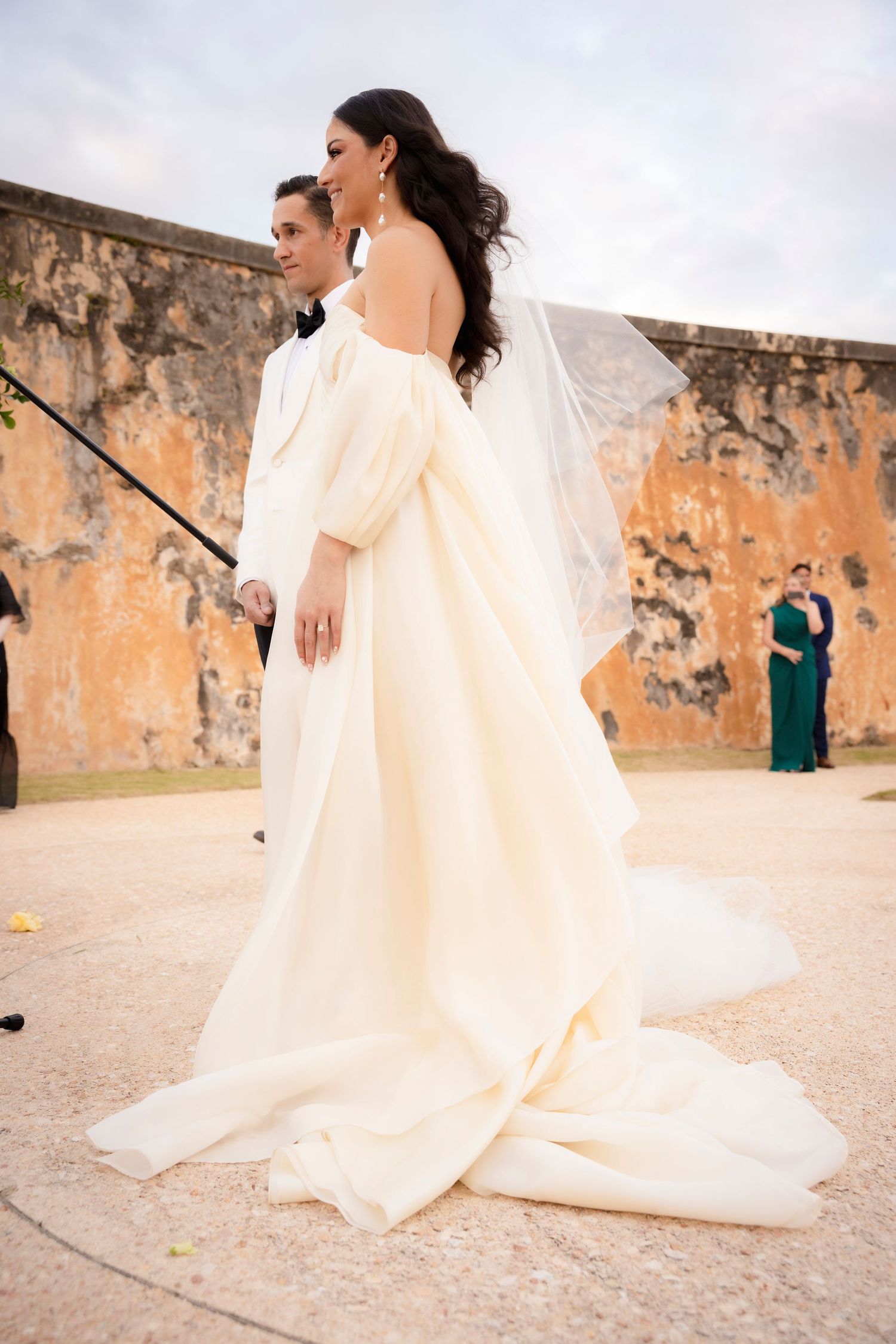 Flowing wedding dress train spreads across stone pathway near historic wall.