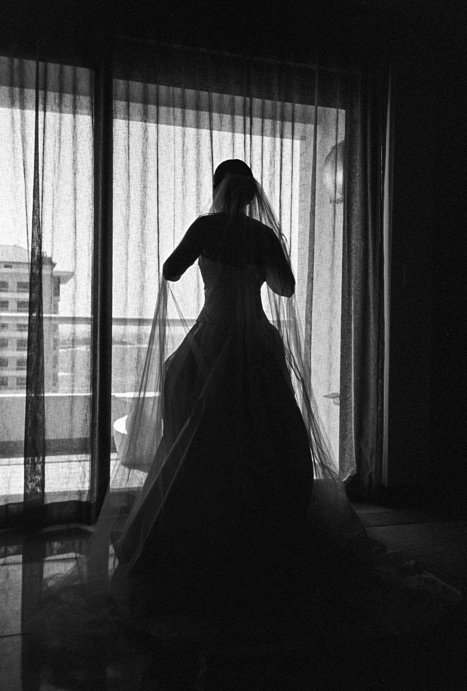 Silhouette of a bride in a flowing wedding gown and veil standing before a large window in black and white.