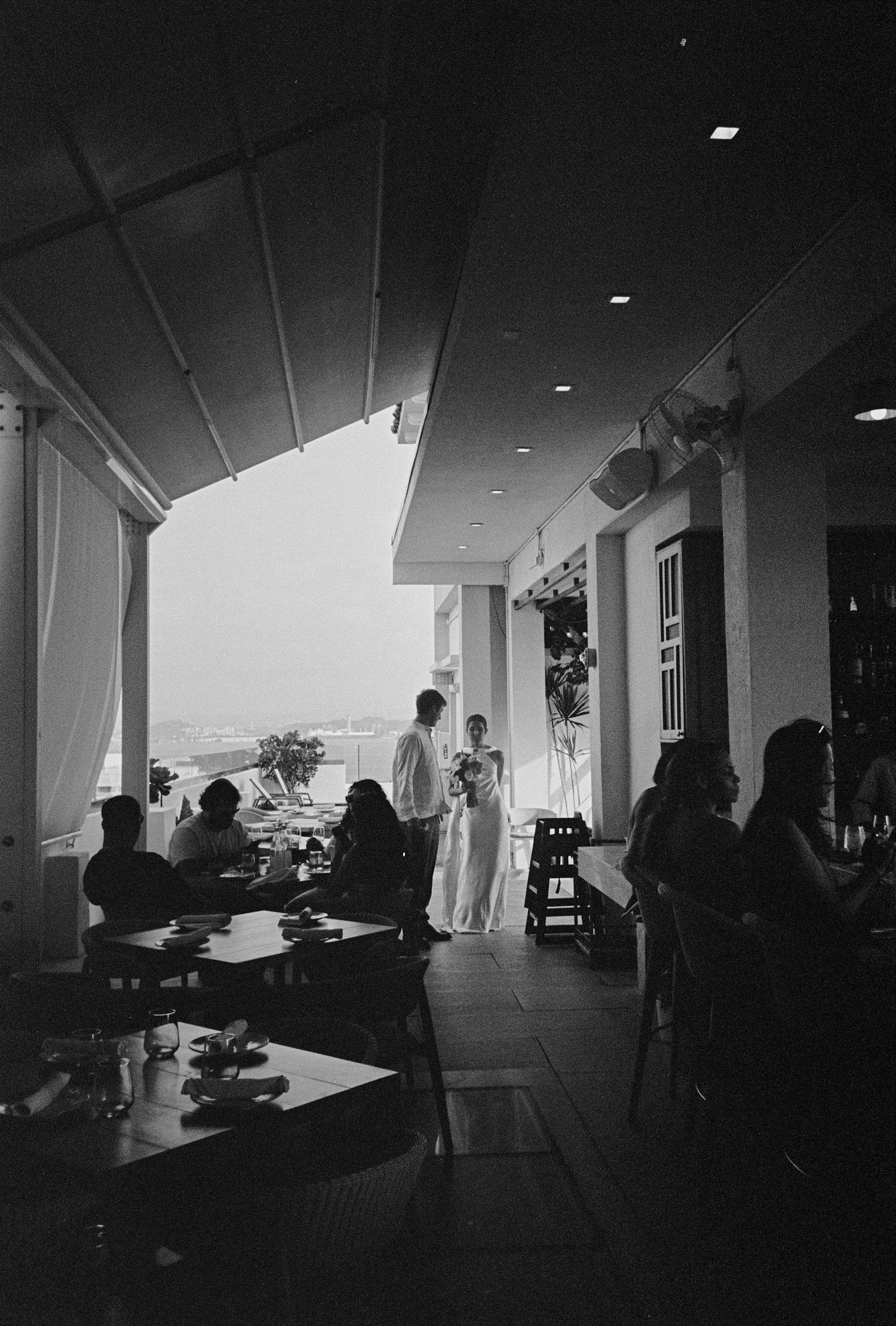 Black and white photo of a wedding couple standing on an elegant restaurant terrace with seated guests.