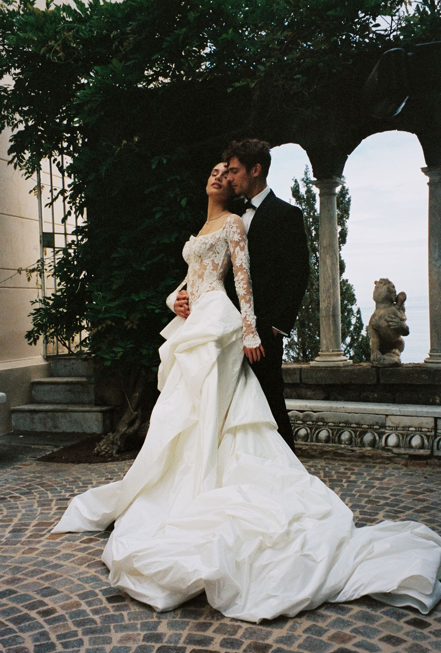Bride and groom pose romantically in a lace long-sleeve wedding gown with dramatic train at a garden venue.