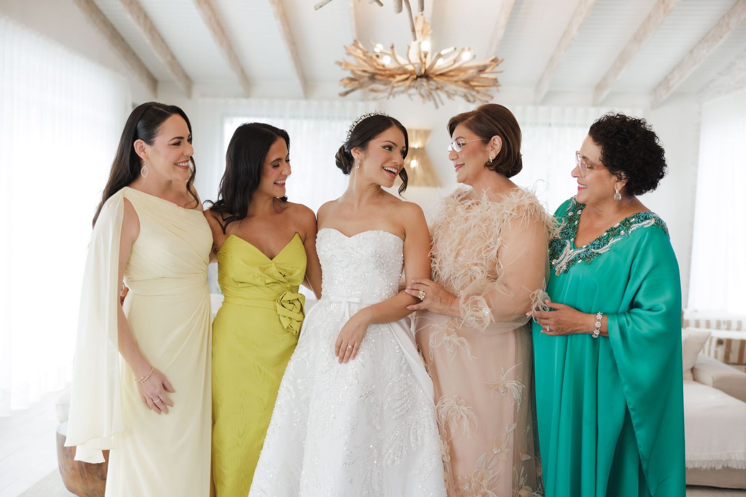 A group in colorful dresses including yellow, white, and turquoise share a joyful moment in a bright indoor space.