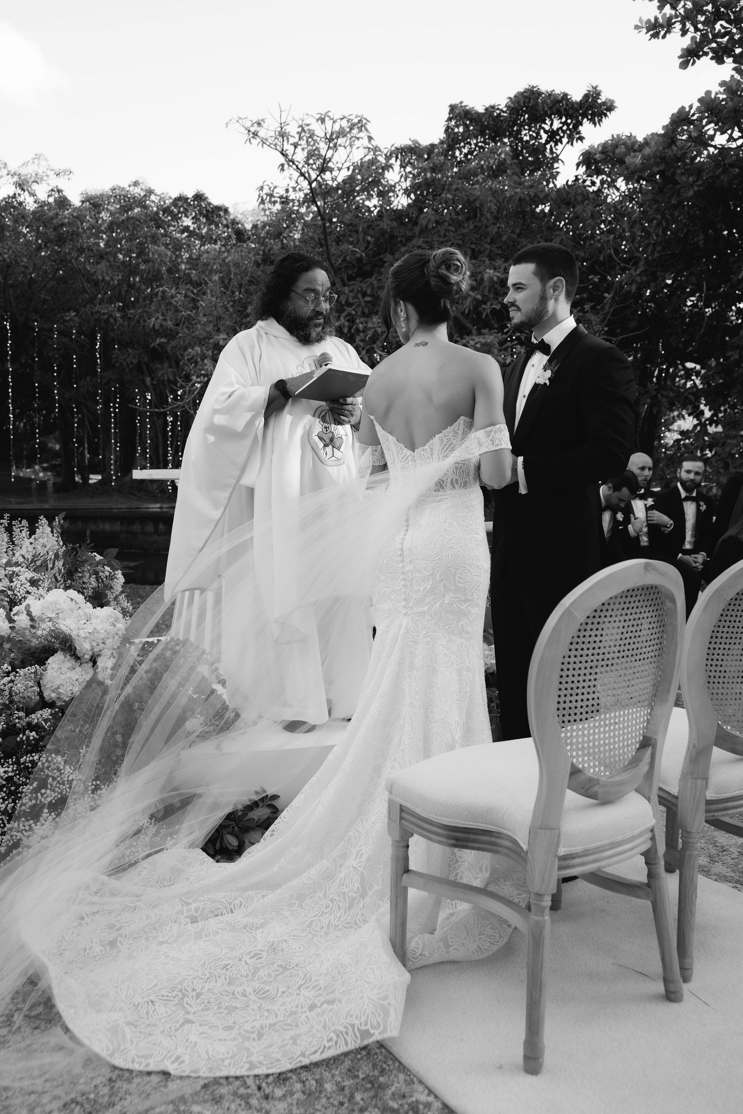 Wedding couple seated on white chairs with flowing bridal train in garden setting.