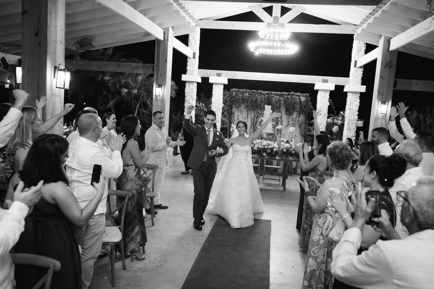 A black and white photo of a wedding reception with guests lining the aisle under string lights.