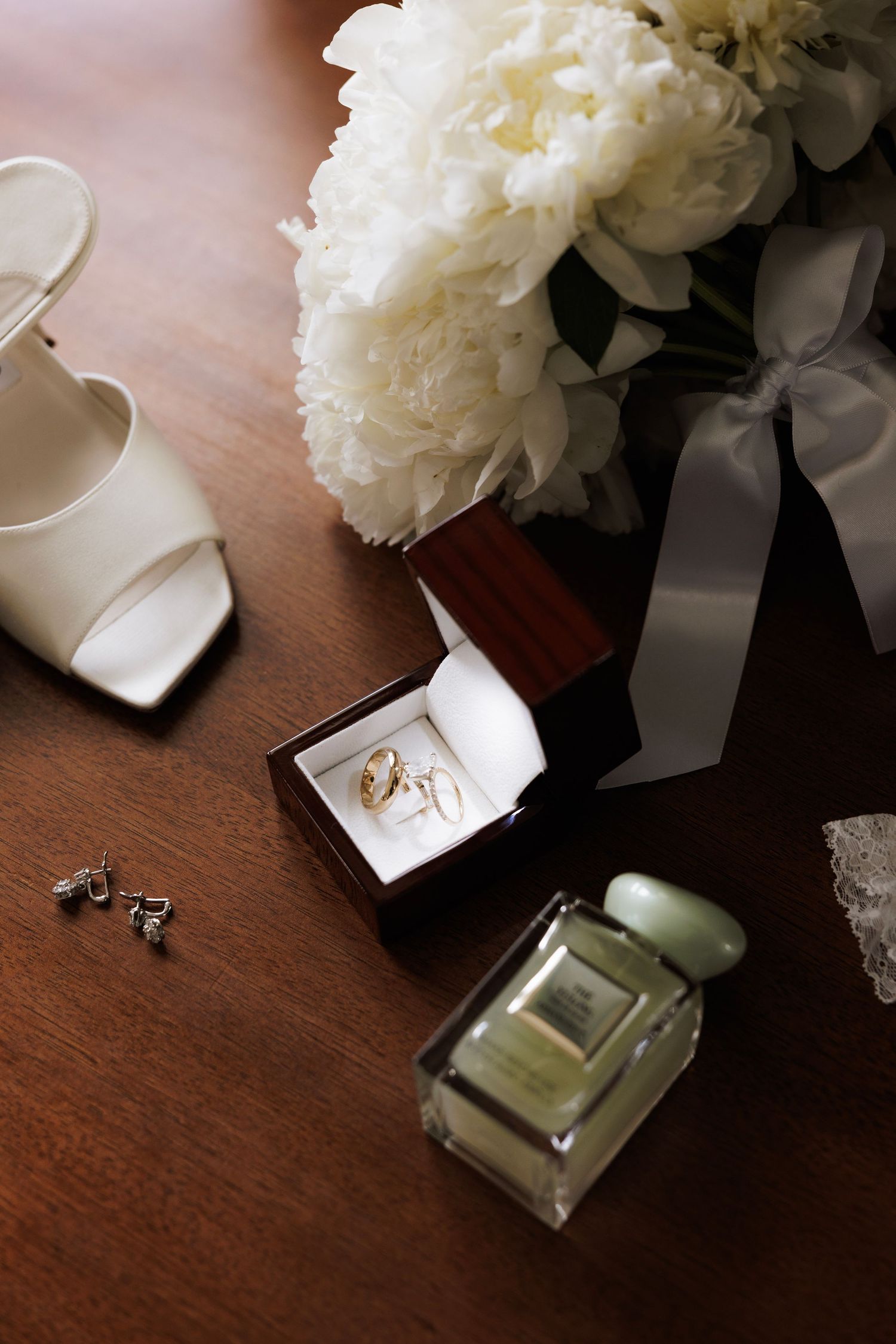 White peonies in a vase next to wedding rings and perfume bottle on dark wooden surface.