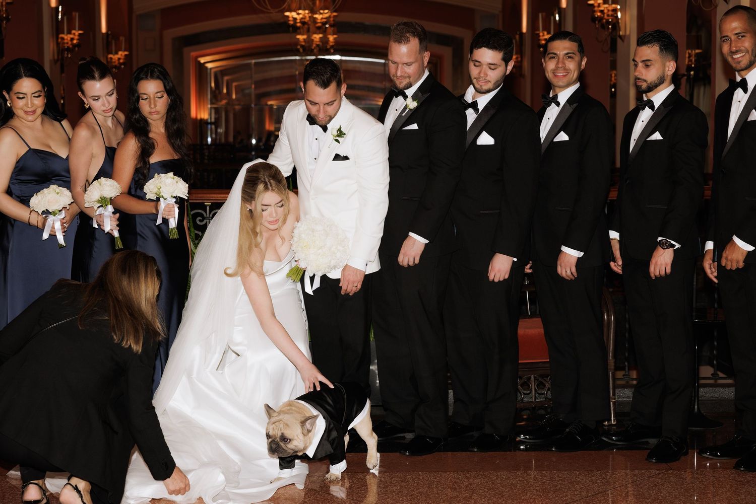 A wedding party stands in formal attire as someone adjusts the bride's dress on a polished ballroom floor.