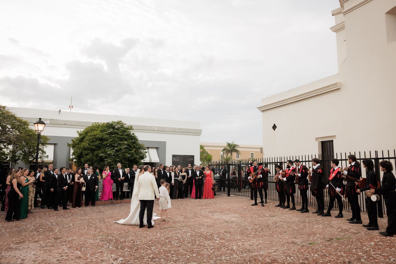 Wedding guests form a receiving line outdoors on a paved walkway with a white building in the background.