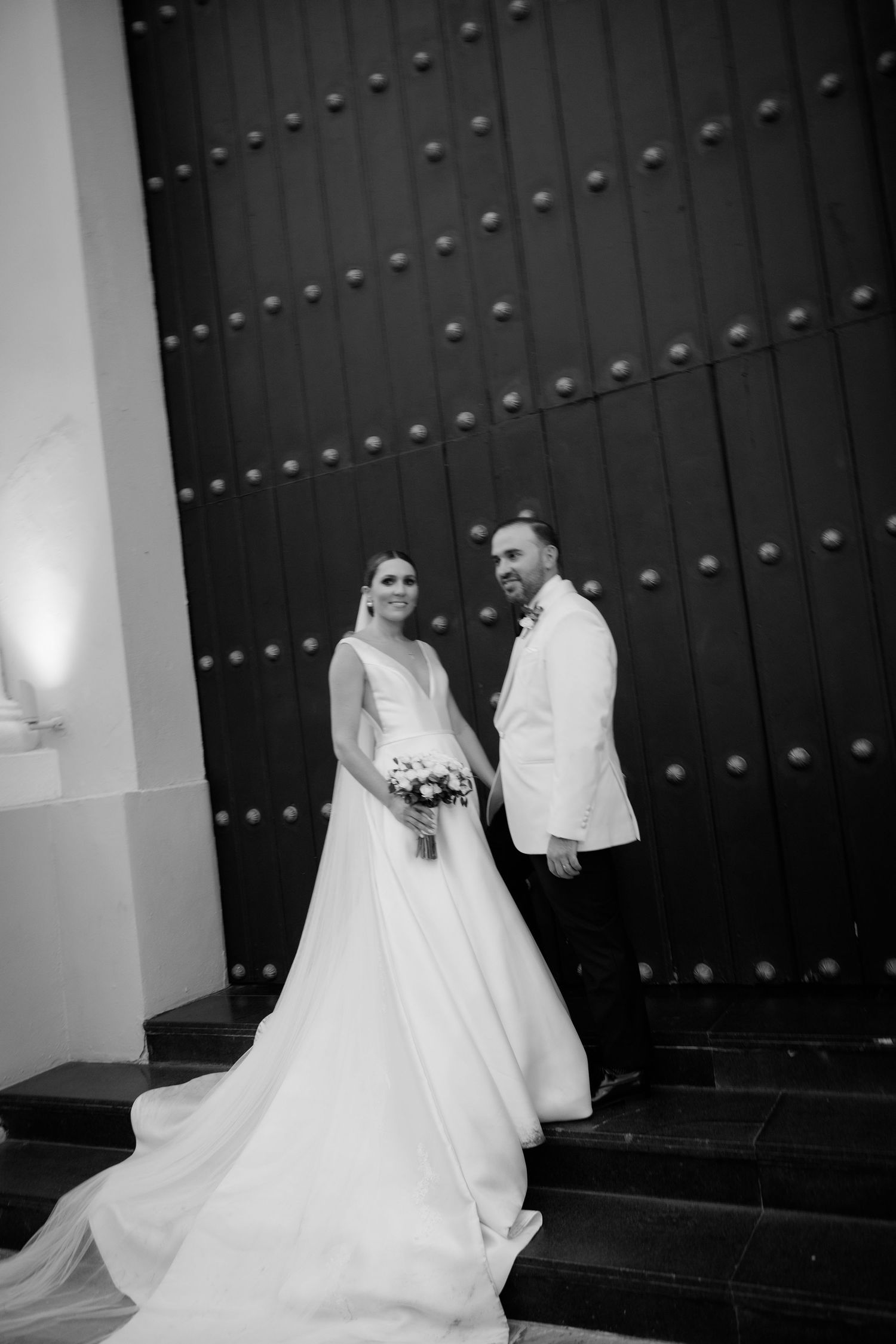 A black and white wedding photo series showing a couple in formal attire posing against dark wooden church doors.