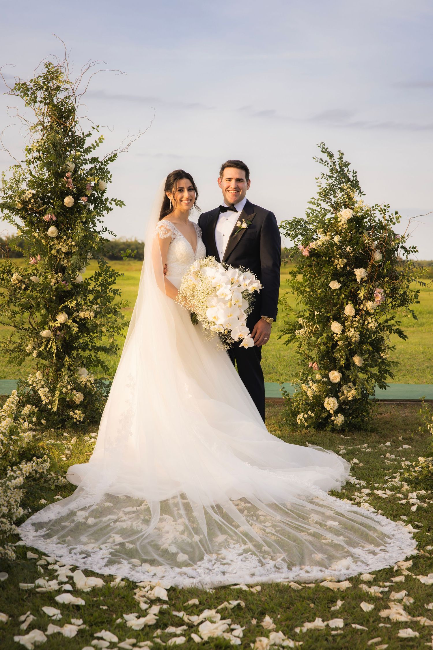 Wedding couple stands in front of lush floral arrangements on a grassy field with scattered white rose petals.