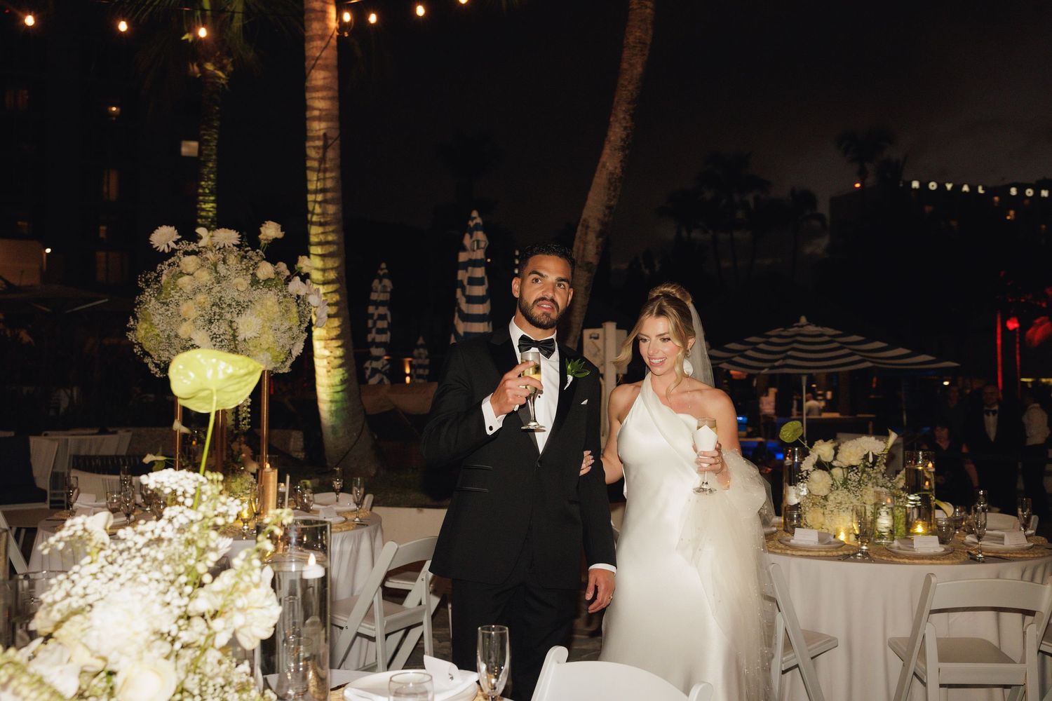 String lights illuminate a nighttime wedding reception with elegant table settings and floral centerpieces.