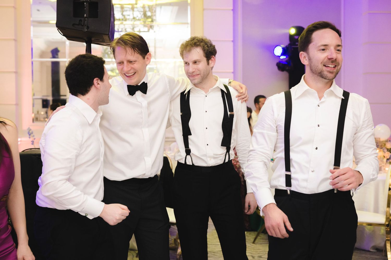 Dancing couples in formal attire enjoy themselves at a wedding reception in an elegant venue with purple lighting.