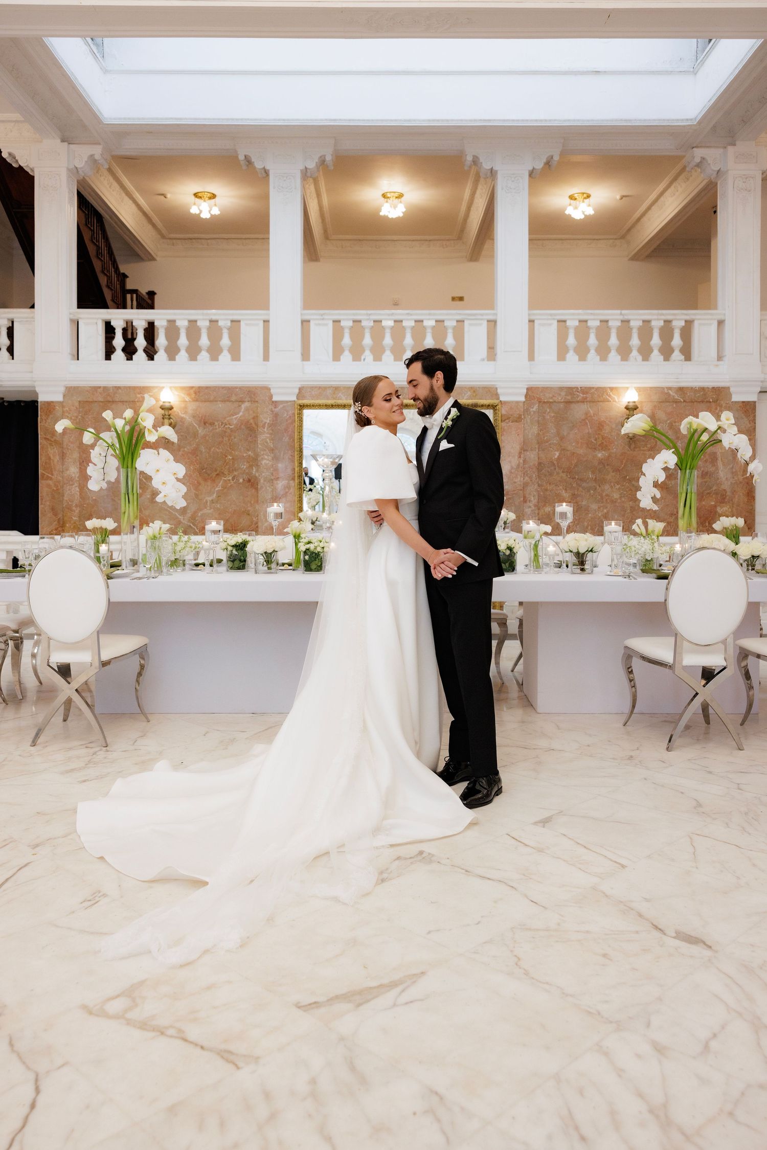 A wedding couple embraces in an elegant marble venue with columns and draped tables in the background.