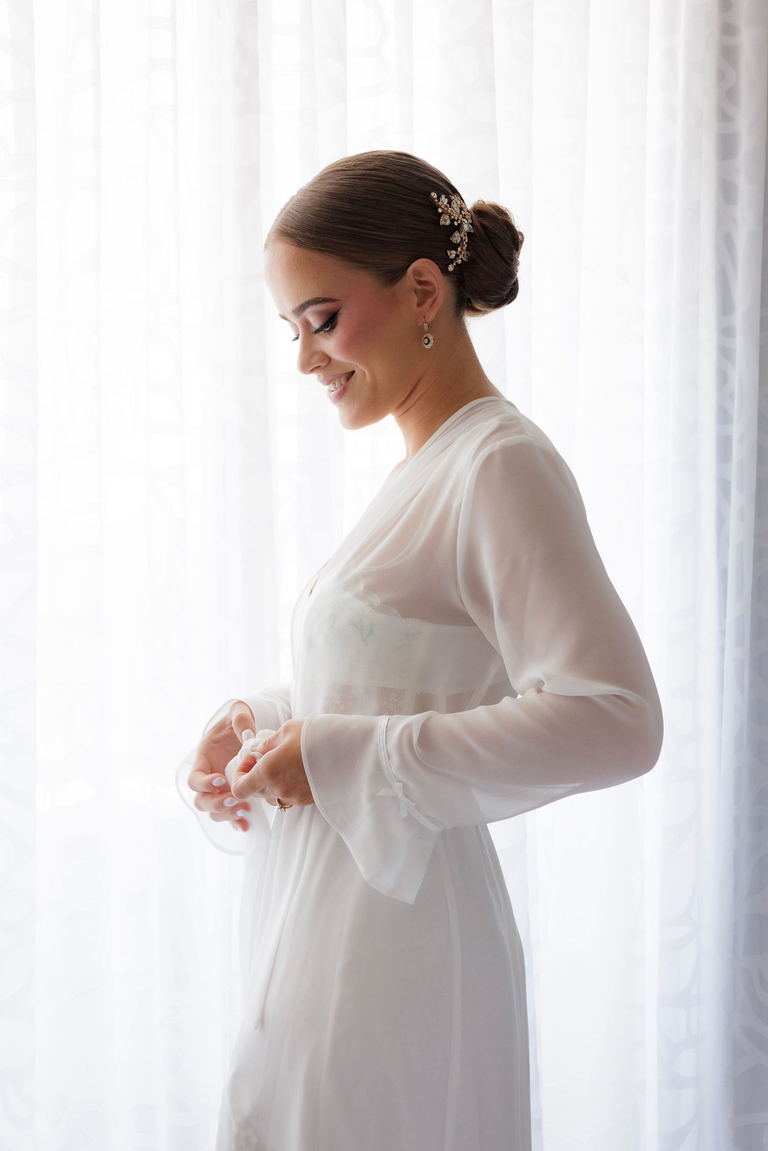 A bride in an elegant sheer sleeve wedding dress poses by a window in soft natural light.