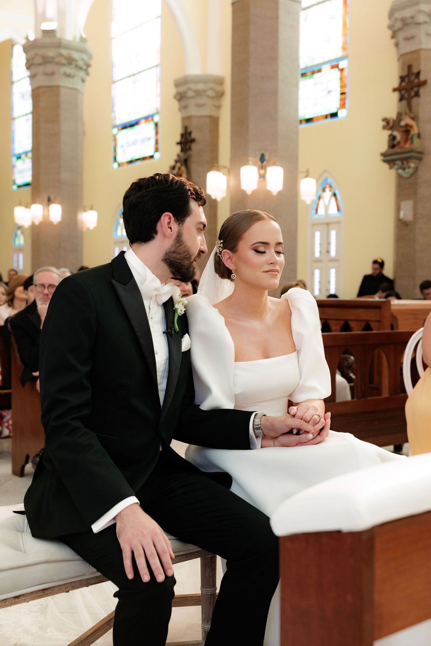 A couple sits together during their wedding ceremony in a traditional church with arched columns and stained glass windows.