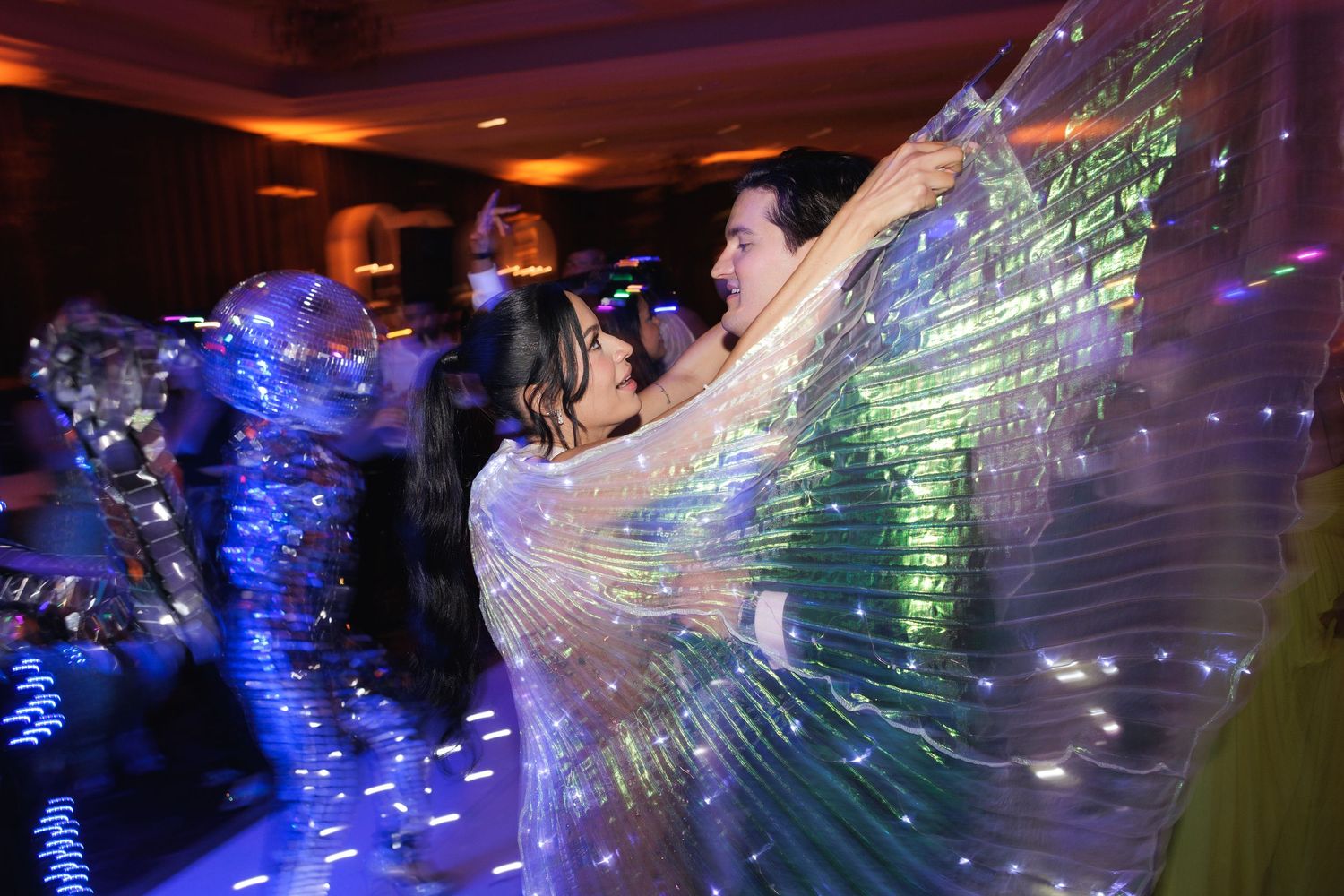Dancers move on a dark dance floor with illuminated flowing fabric wings creating streaks of blue and green light effects.