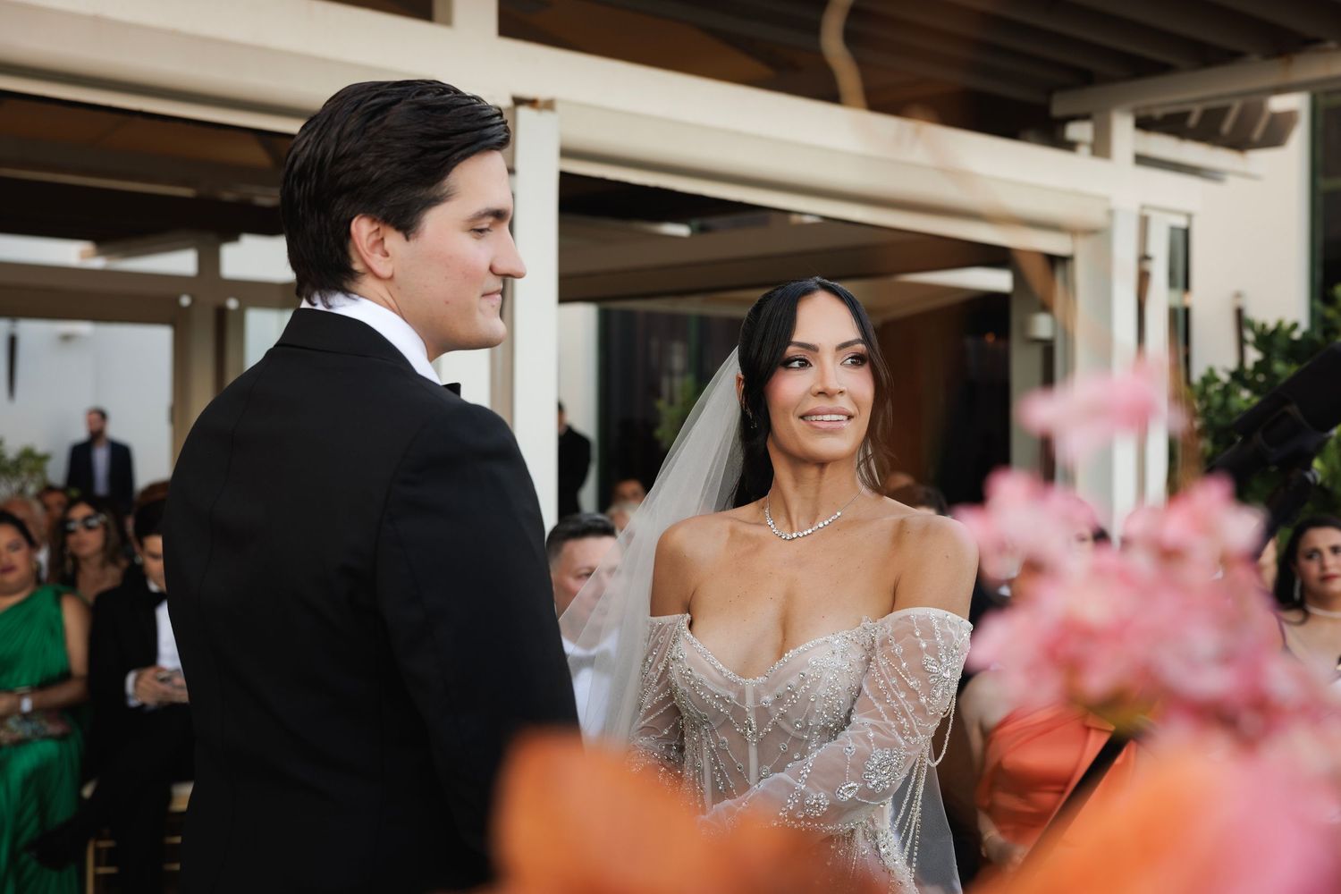 Couple shares an intimate moment during wedding ceremony with pink floral decor in foreground.