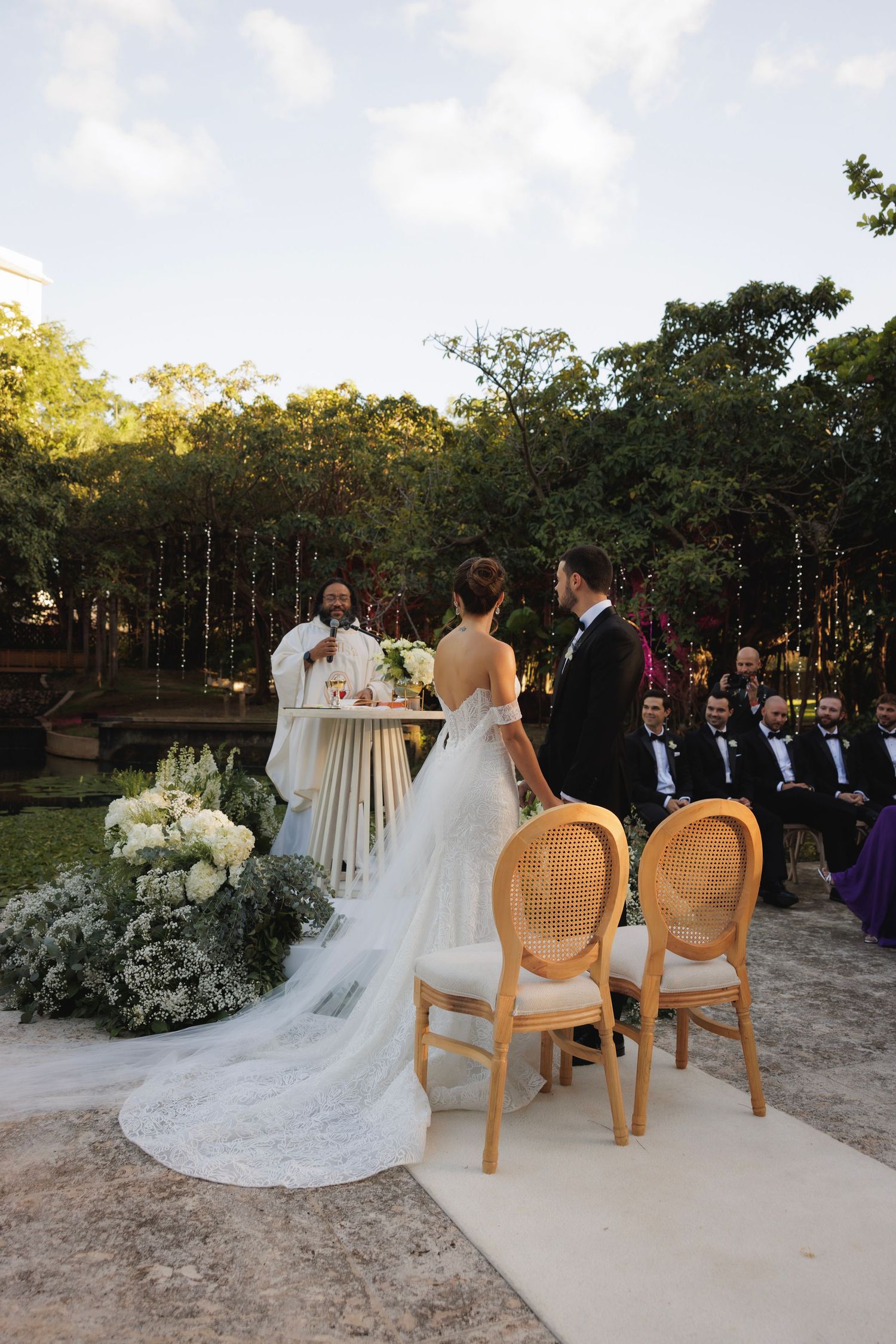 Garden wedding ceremony with vintage cane chairs and abundant white flowers against a backdrop of tall trees.