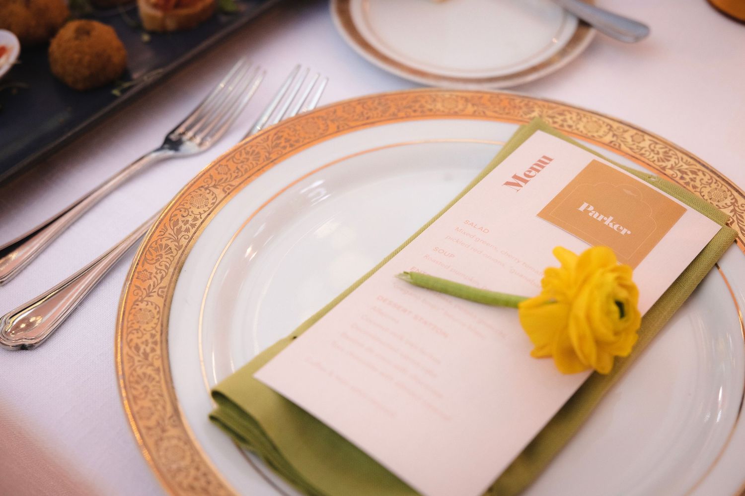 An elegant place setting with white dinnerware, gold charger plate, and yellow daffodil accent at a wedding reception.