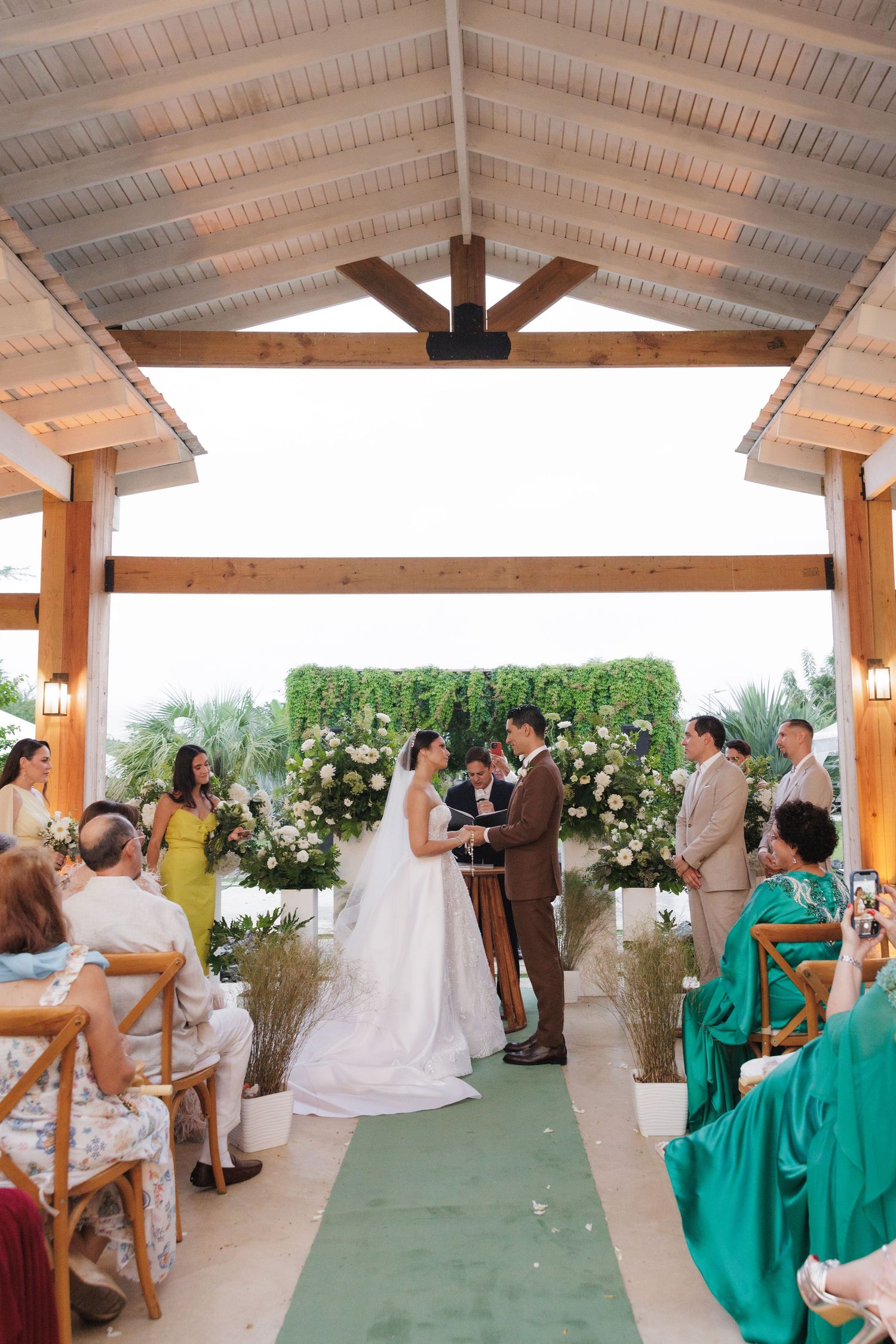 Wedding ceremony setup under a covered outdoor pavilion with green bridesmaid dresses and white floral decorations.