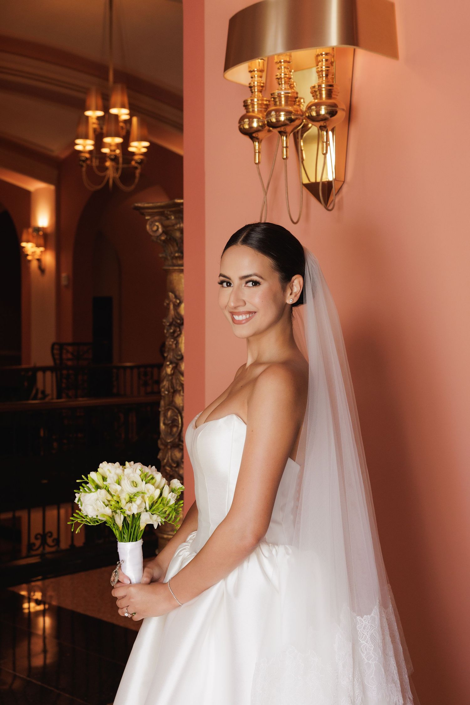 Bride in classic strapless gown poses with bouquet under elegant chandelier lighting.