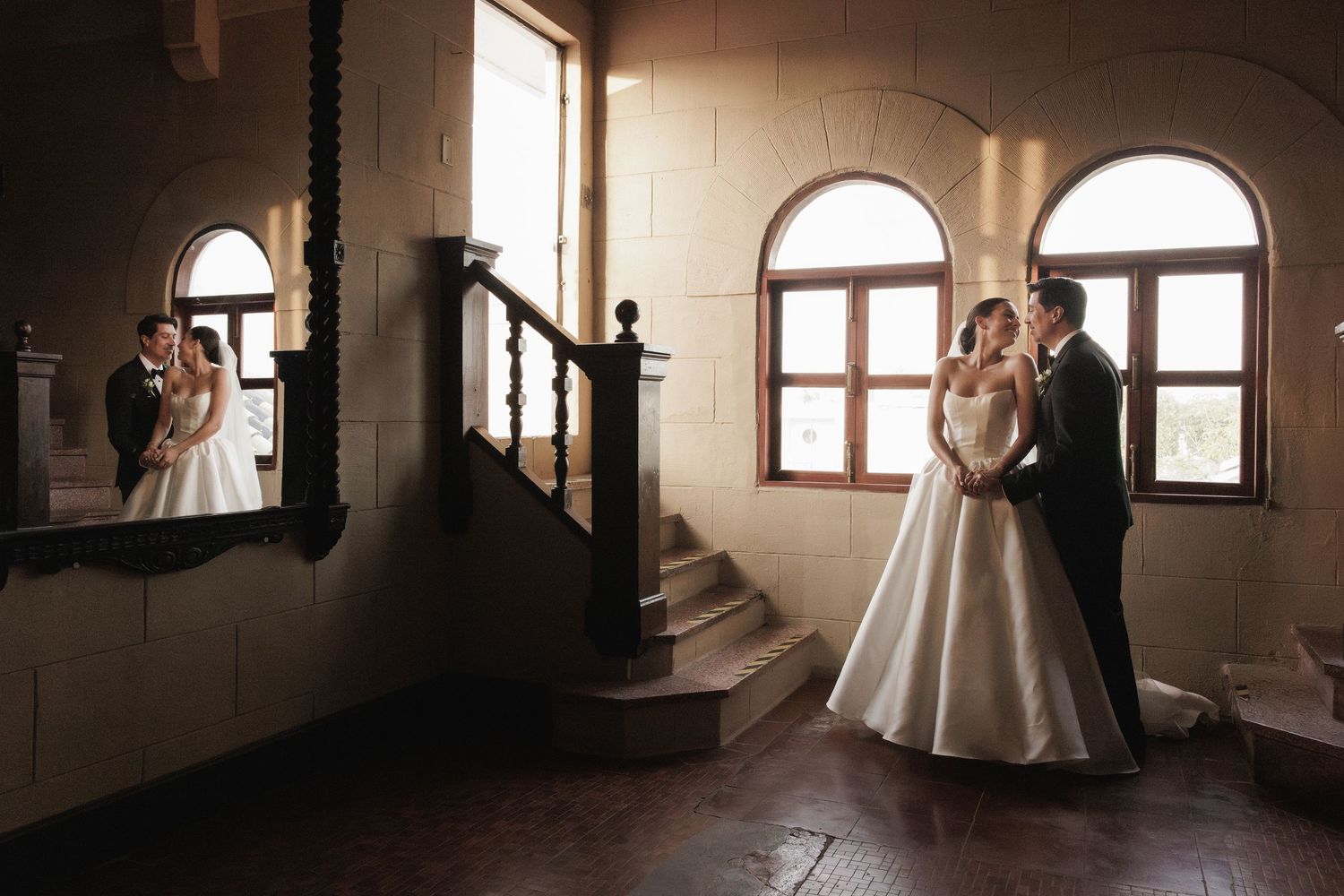 A bride in a flowing white gown poses near sunlit windows in a dark rustic interior space.
