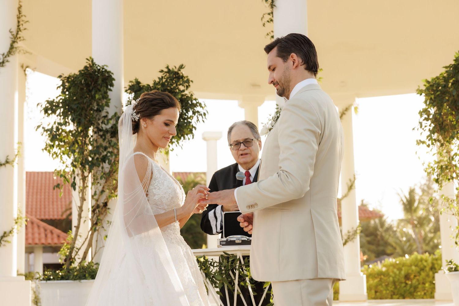 A couple exchanges rings during an elegant outdoor wedding ceremony beneath a gazebo with decorative greenery.